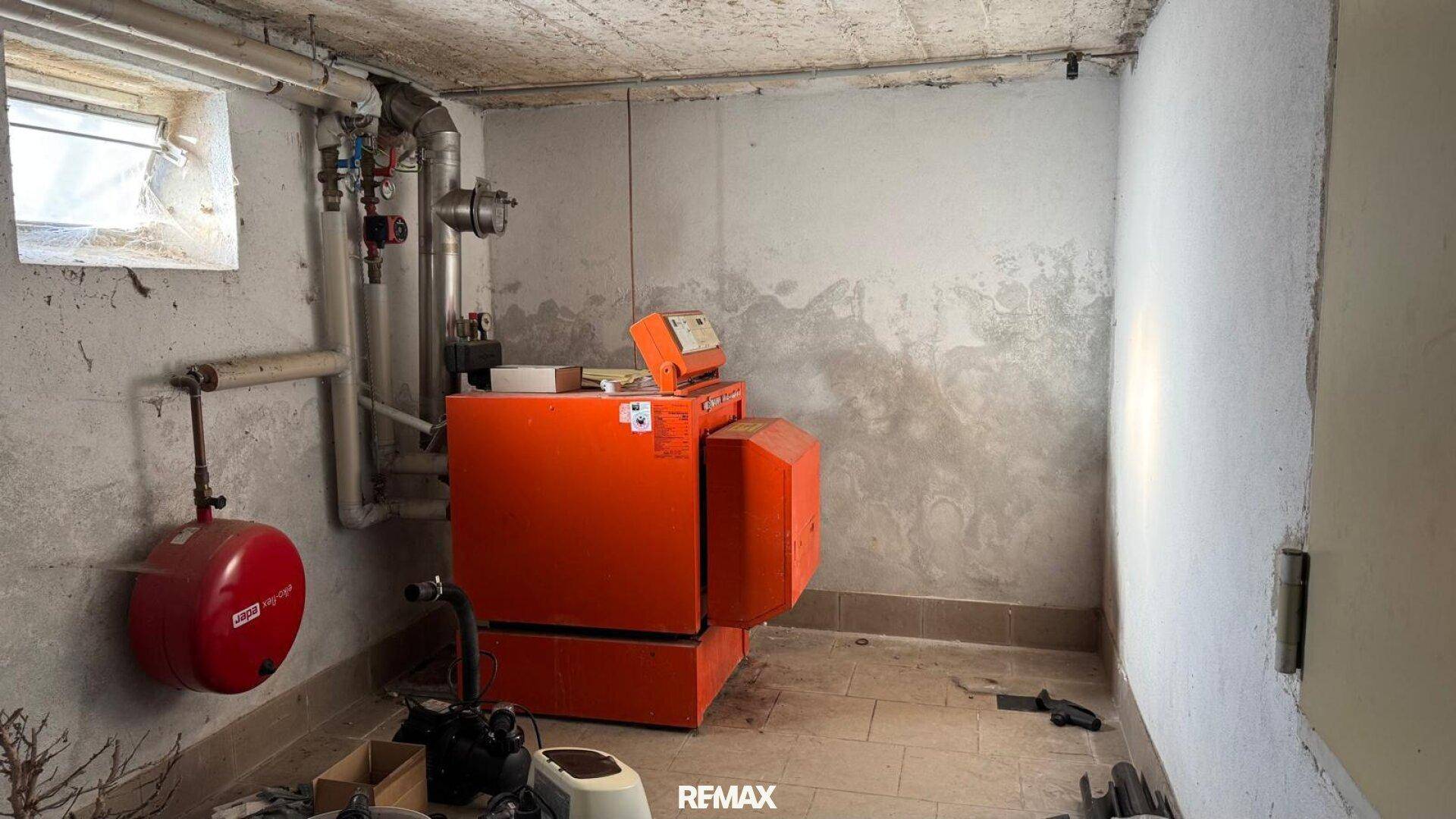 Heizungsraum im Keller mit einem großen orangefarbenen Heizkessel und einem kleinen Fenster.