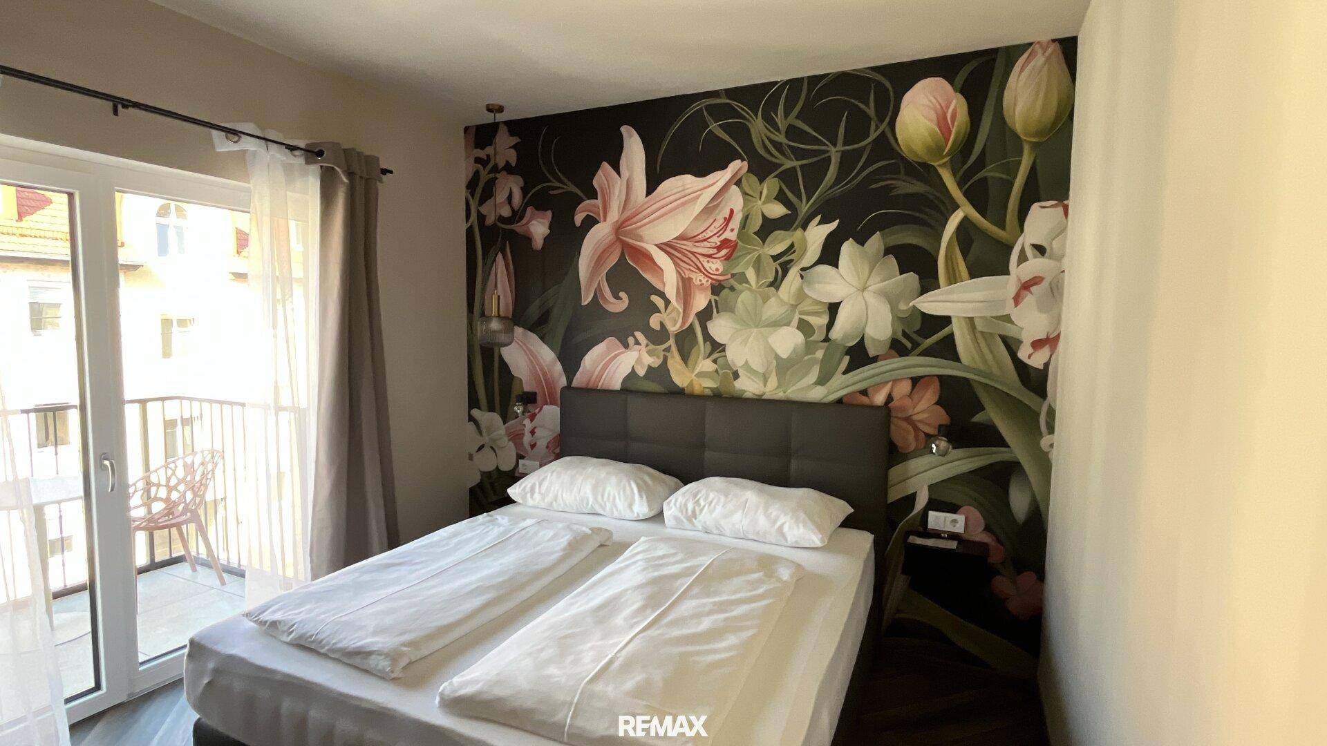 Helles Schlafzimmer mit Doppelbett, floraler Wandtapete und Zugang zu einem Balkon.