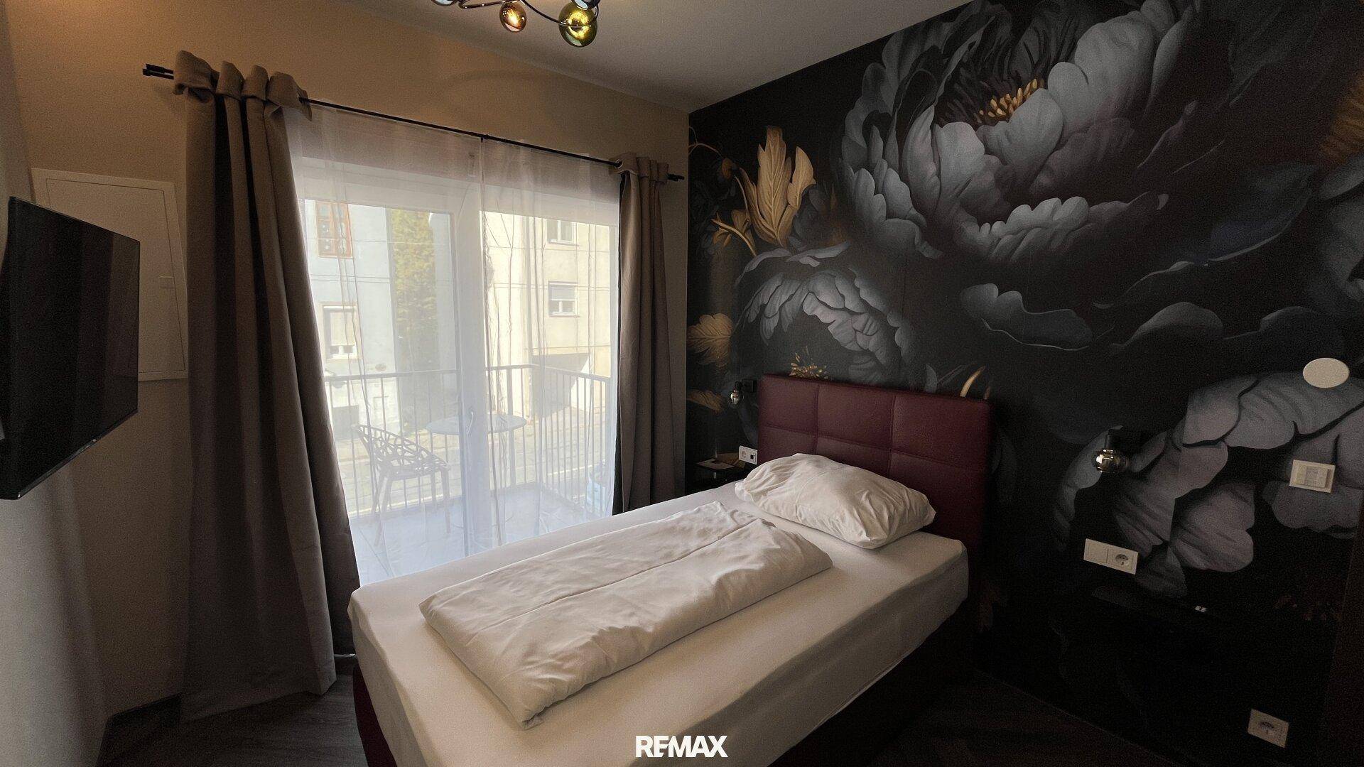 Schlafzimmer mit Einzelbett, dunkler floraler Wandtapete und Balkonzugang.