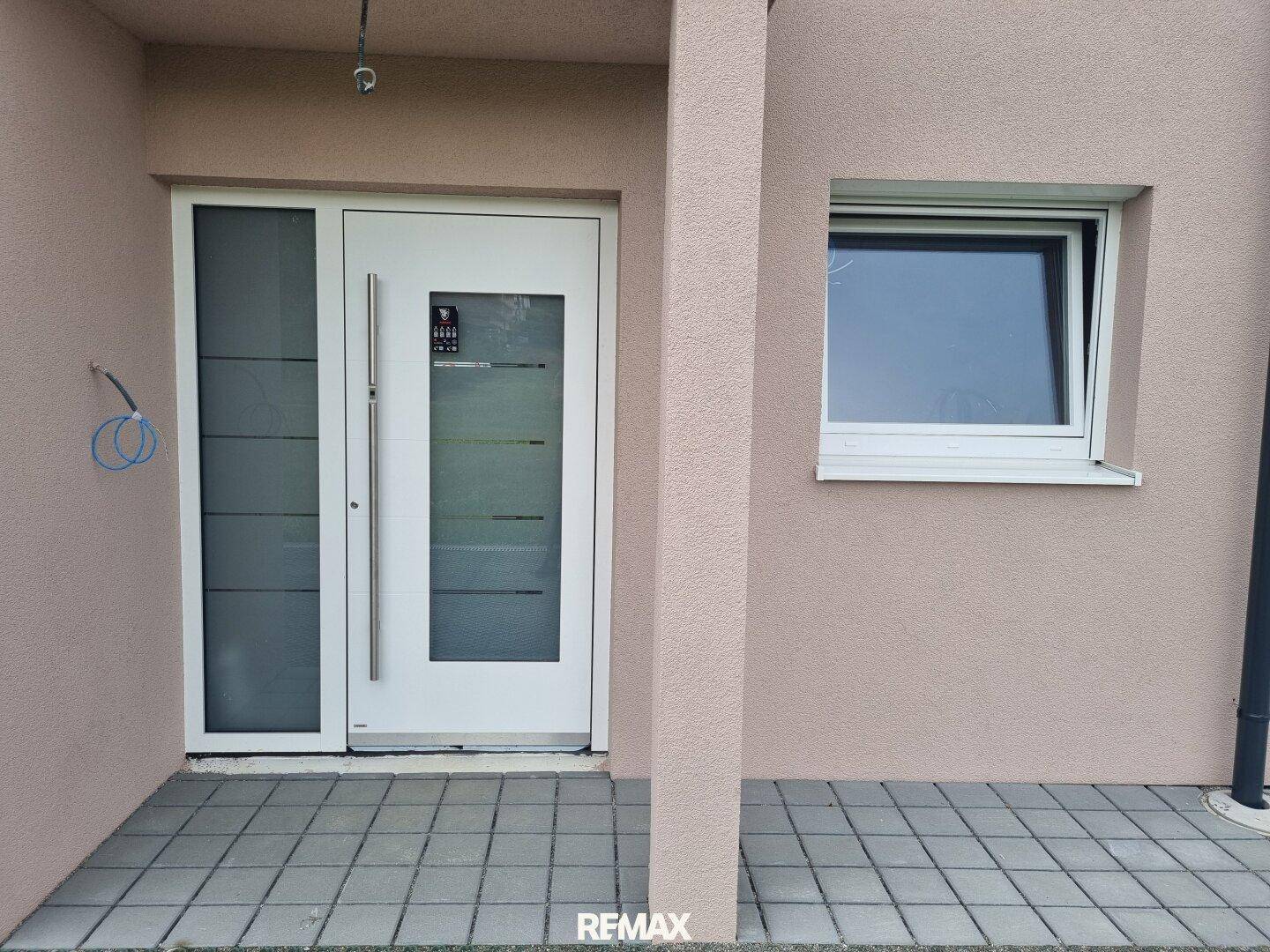 Moderner Hauseingang mit weißer Tür und seitlichem Fenster, umgeben von beigefarbener Fassade.