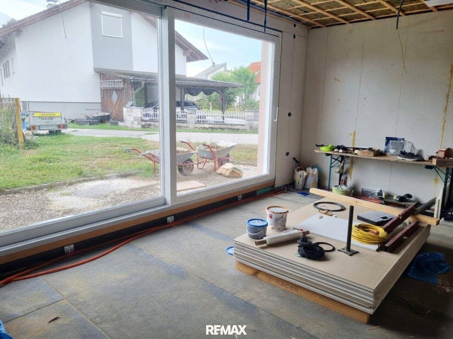 Innenraum im Rohbau mit großer Fensterfront und Blick auf den Garten, mit Baumaterialien.