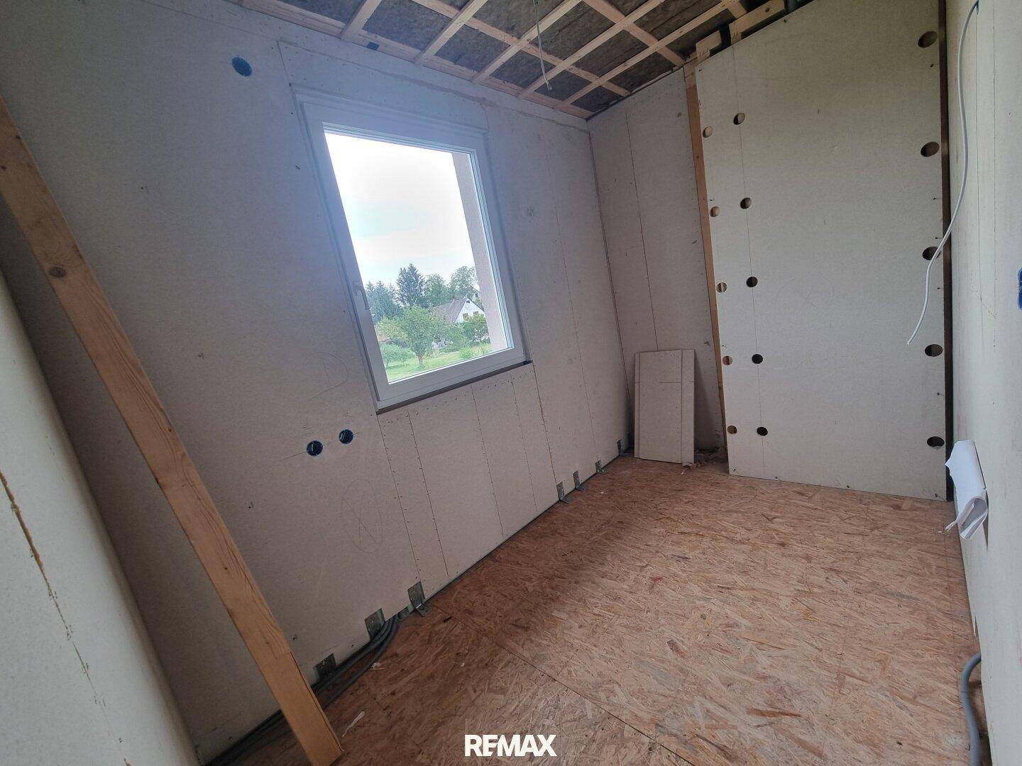 Rohbauzimmer mit einem Fenster und Holzspanplattenboden, bereit für den Innenausbau.