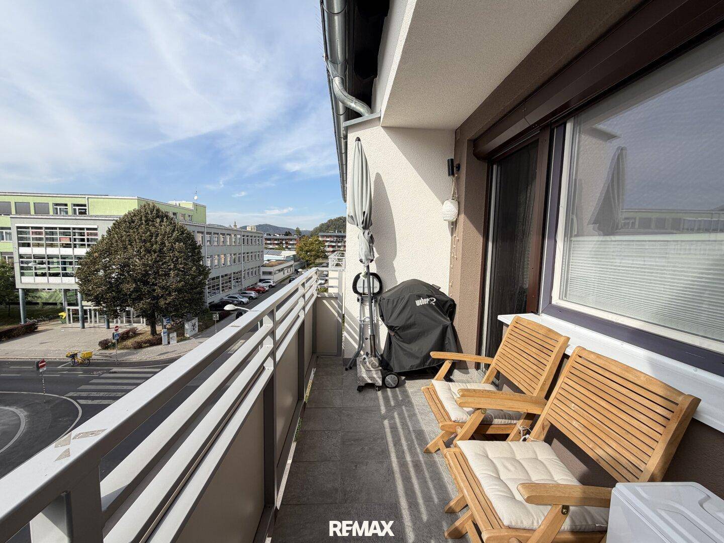 Schmaler Balkon mit Holzsitzmöbeln, Grill und Blick auf die umliegende Stadtlandschaft.
