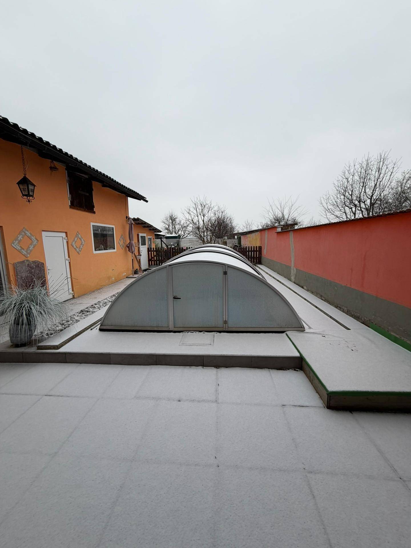 Seitenansicht des Hauses mit orangefarbener Fassade, schneebedecktem Weg und einer überdachten Poolstruktur im Garten.