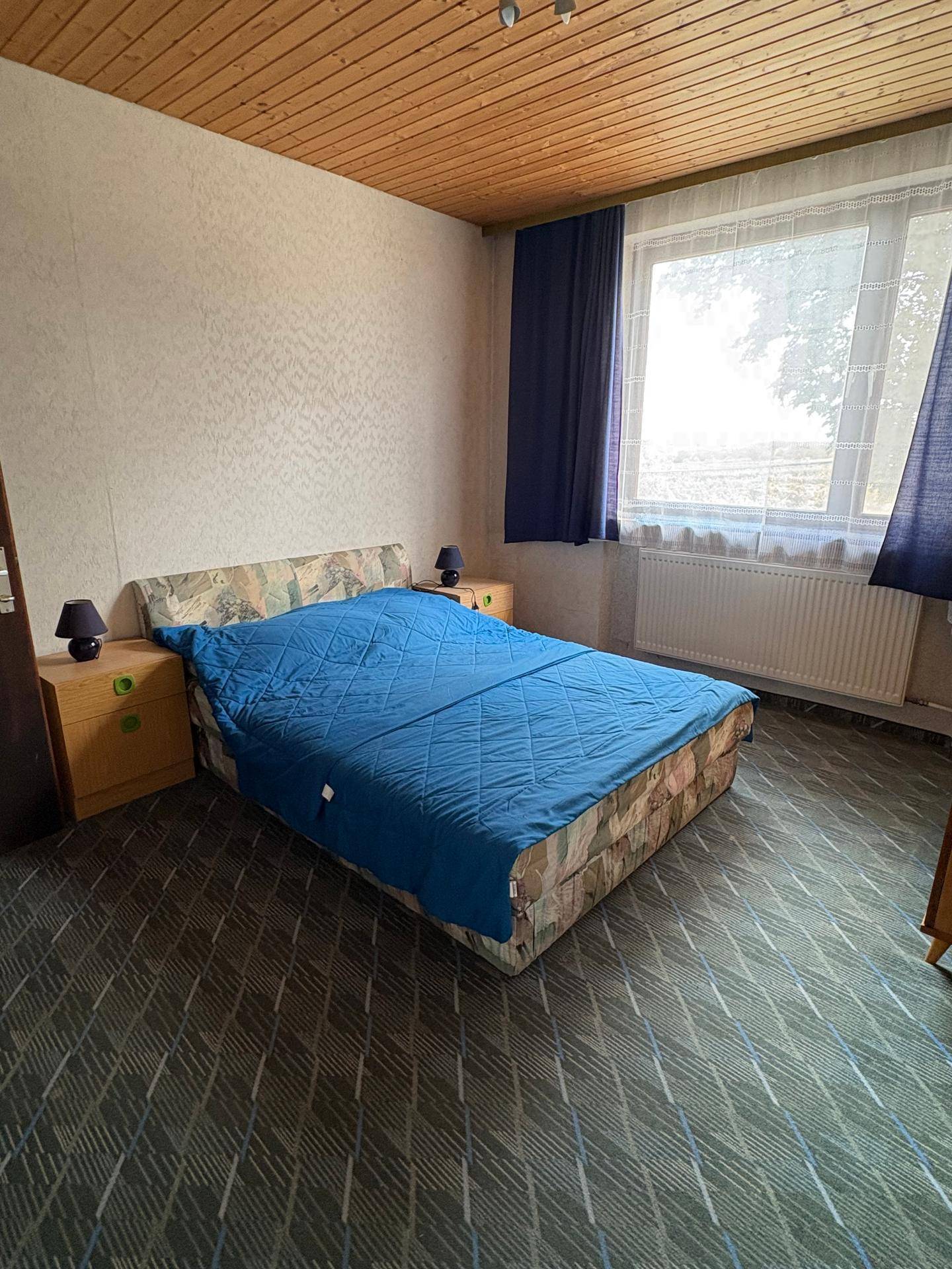Schlafzimmer mit Doppelbett, Teppichboden und großem Fenster mit blauen Vorhängen.