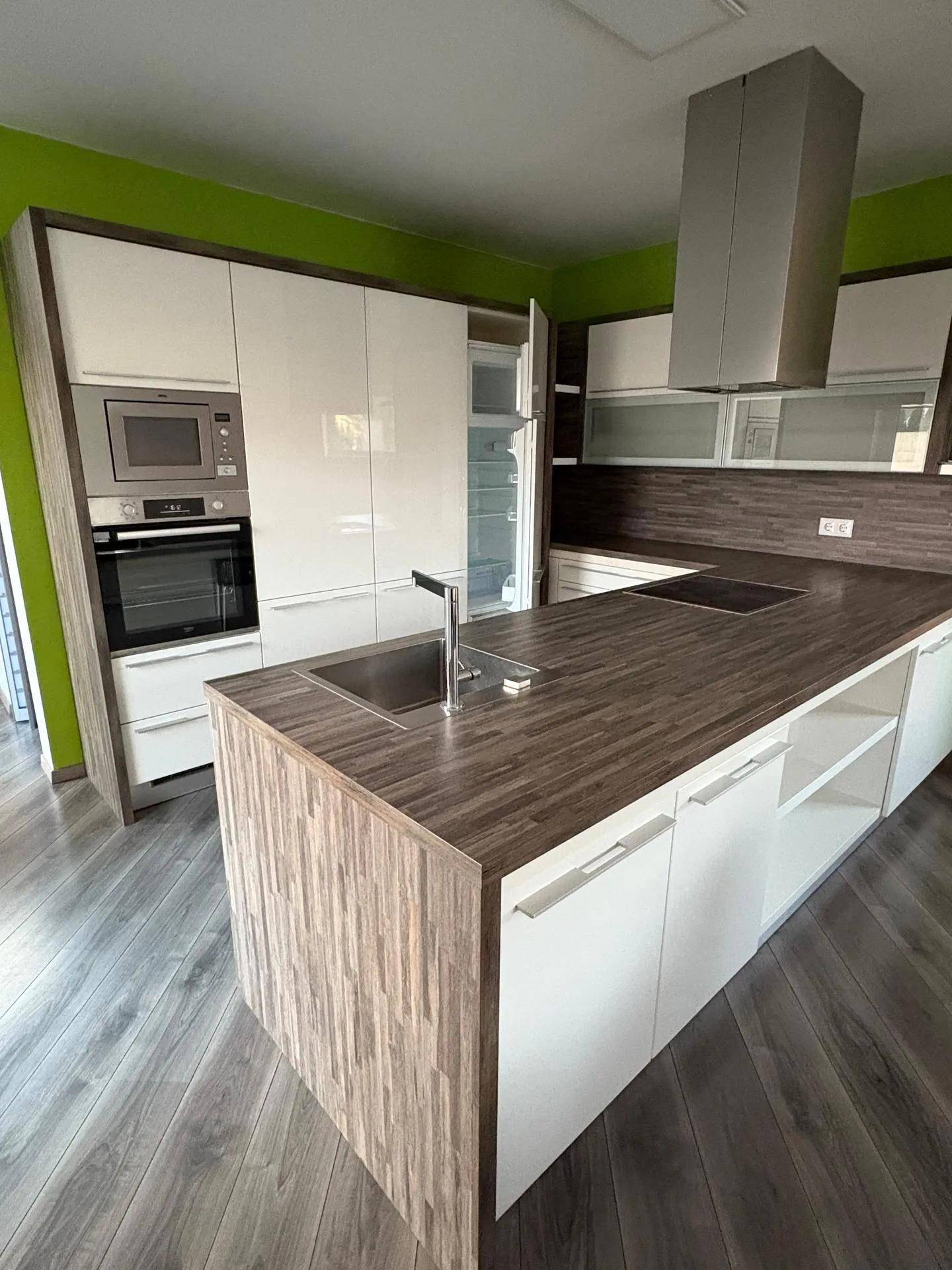 Moderne Einbauküche mit Kochinsel, weißen Hochglanzfronten und Holzarbeitsplatten in einem hellen Raum.