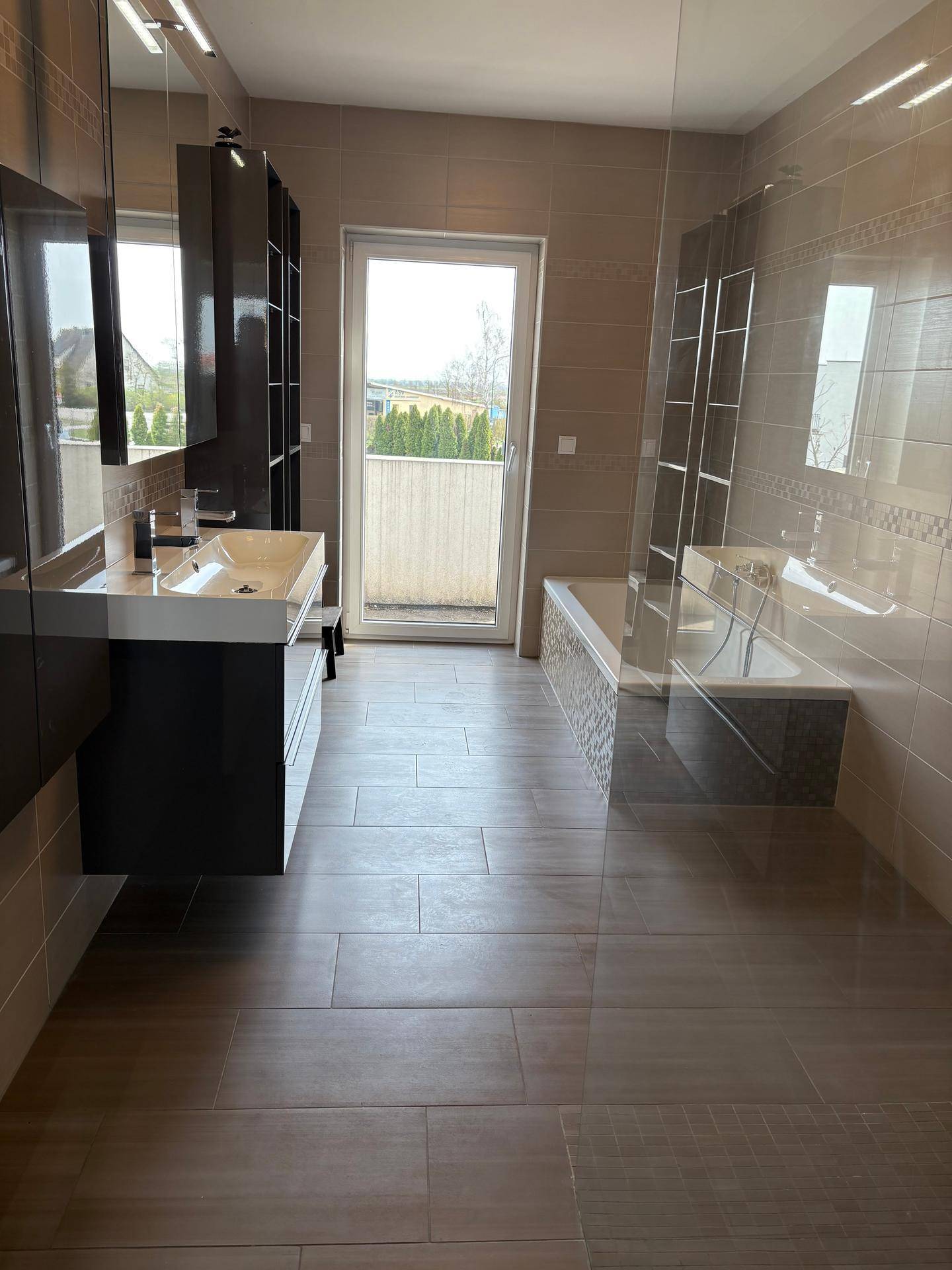 Modernes Badezimmer mit beigefarbenen Fliesen, Doppelwaschbecken und Badewanne mit Zugang zum Balkon.