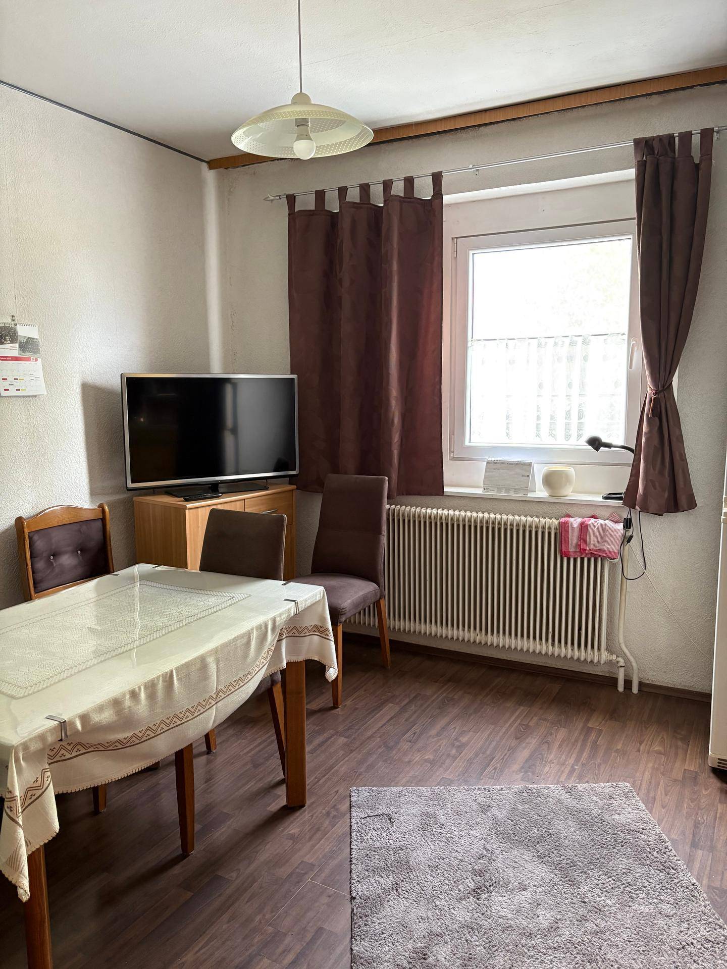 Wohn-/Essbereich mit Fernseher, Esstisch, dunklem Laminatboden und Fenster mit braunen Vorhängen.