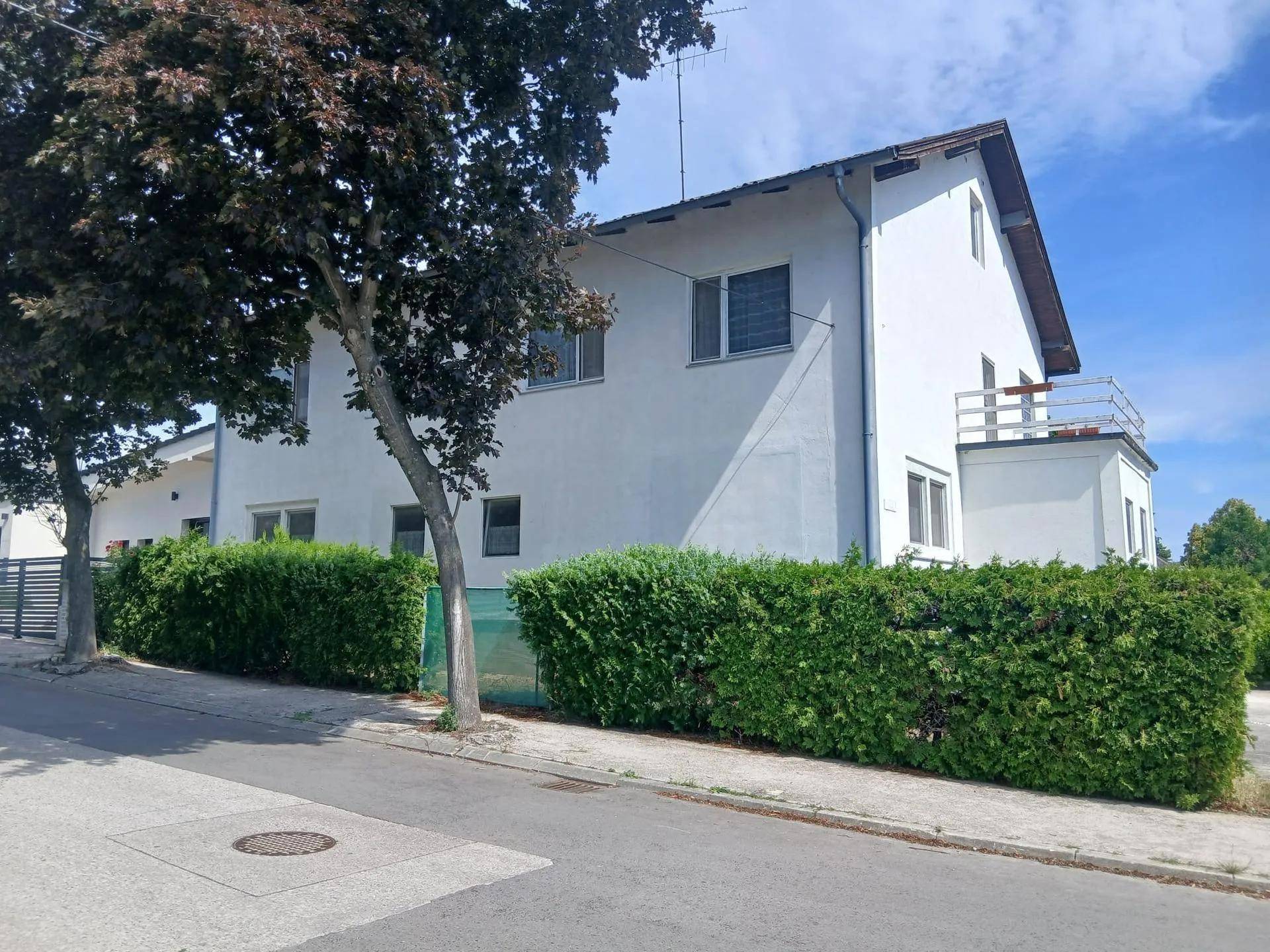 Weißes Wohnhaus mit Hecke und Bäumen an einer ruhigen Straße.