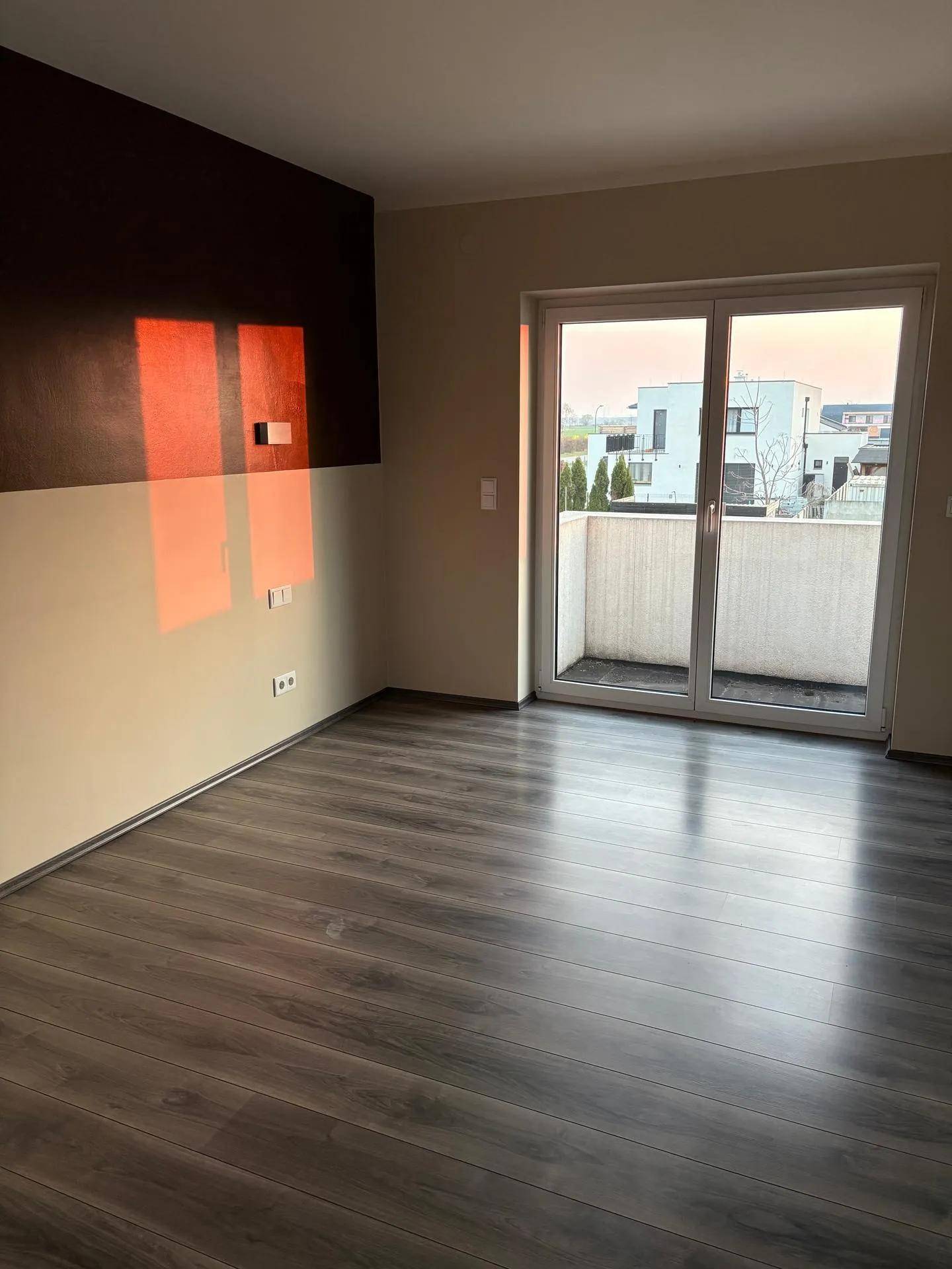 Geräumiges Zimmer mit dunklem Laminatboden und Zugang zu einem Balkon, beleuchtet von der Abendsonne.