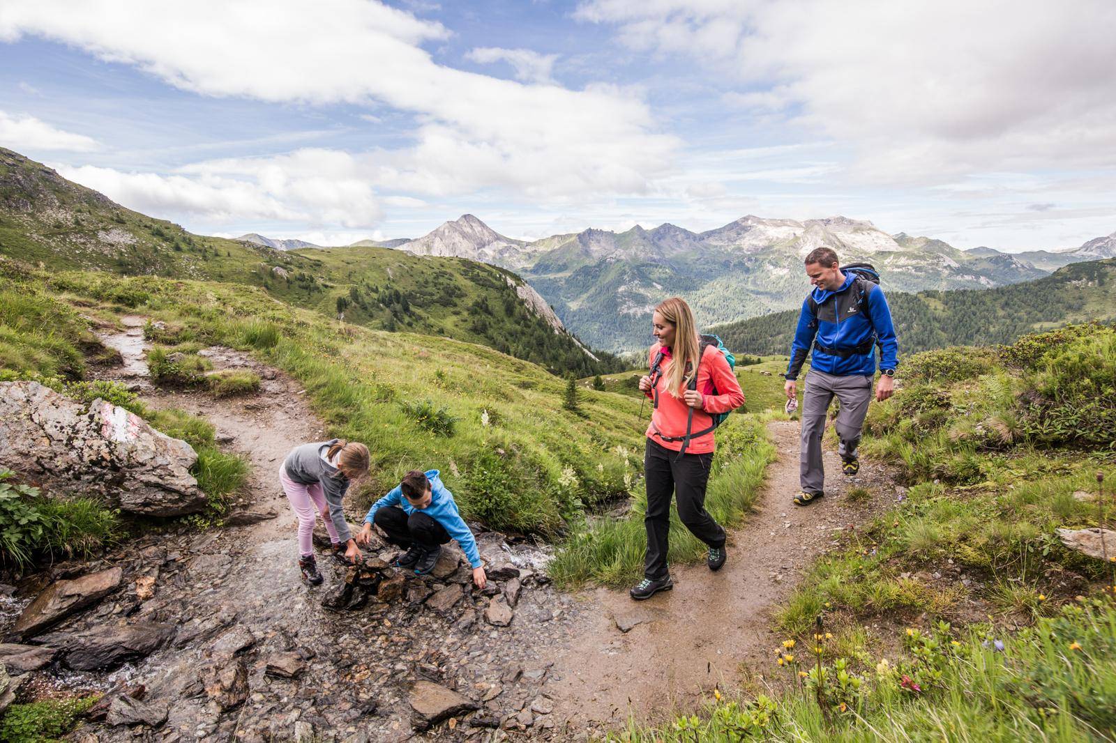 Familie wandert in den Bergen, Kinder spielen an einem Bachlauf, umgeben von grüner Natur.