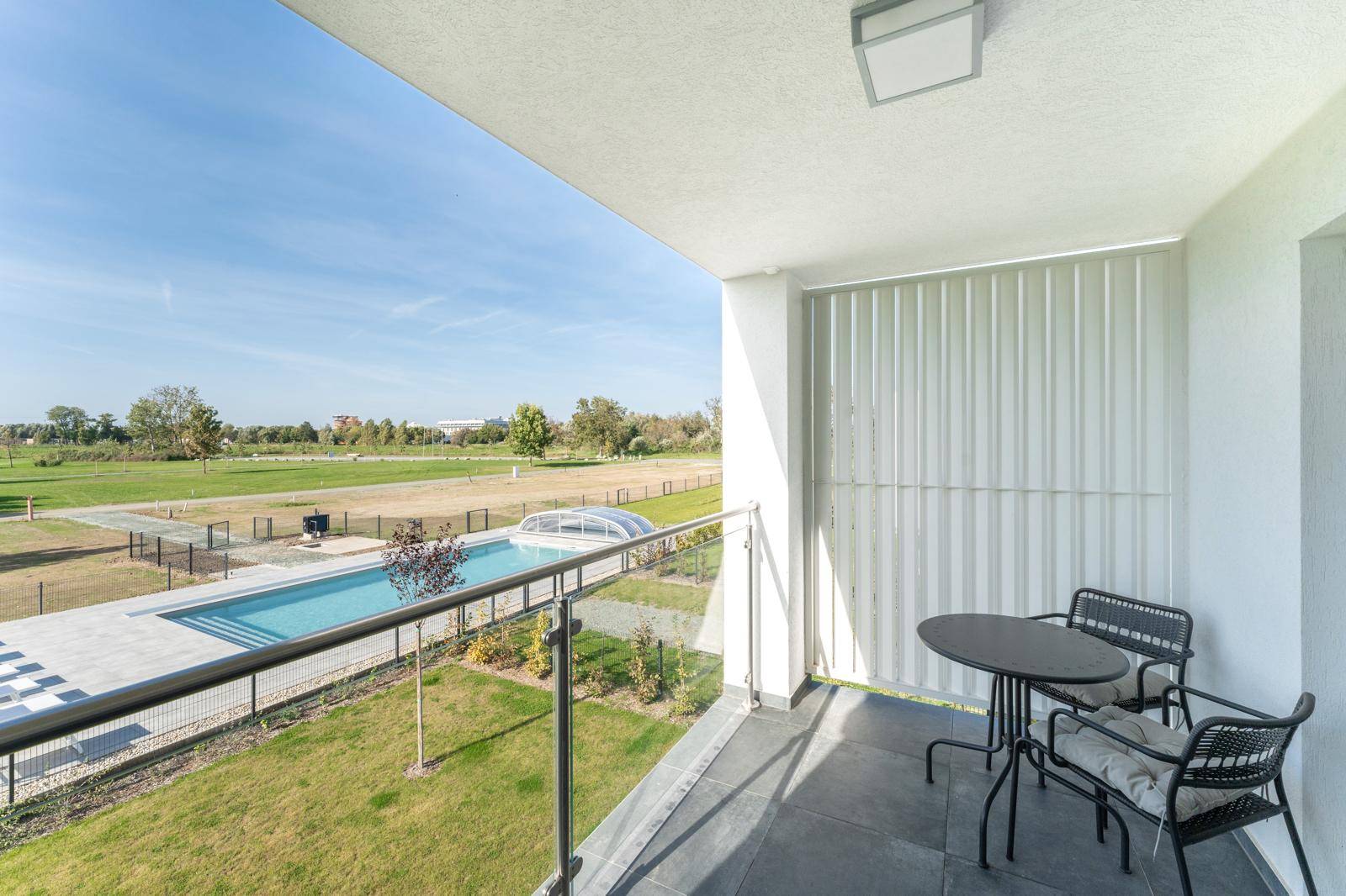 Heller Balkon mit modernen Gartenmöbeln und weitem Blick auf eine Grünfläche mit Swimmingpool.