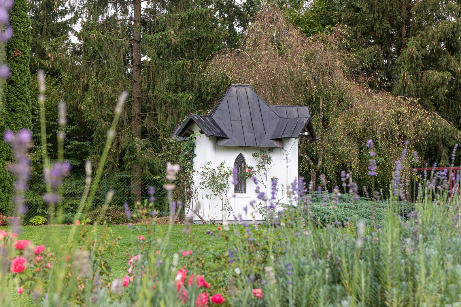 Charmantes kleines Gartenhaus mit besonderem Dach, umgeben von blühenden Blumenbeeten.