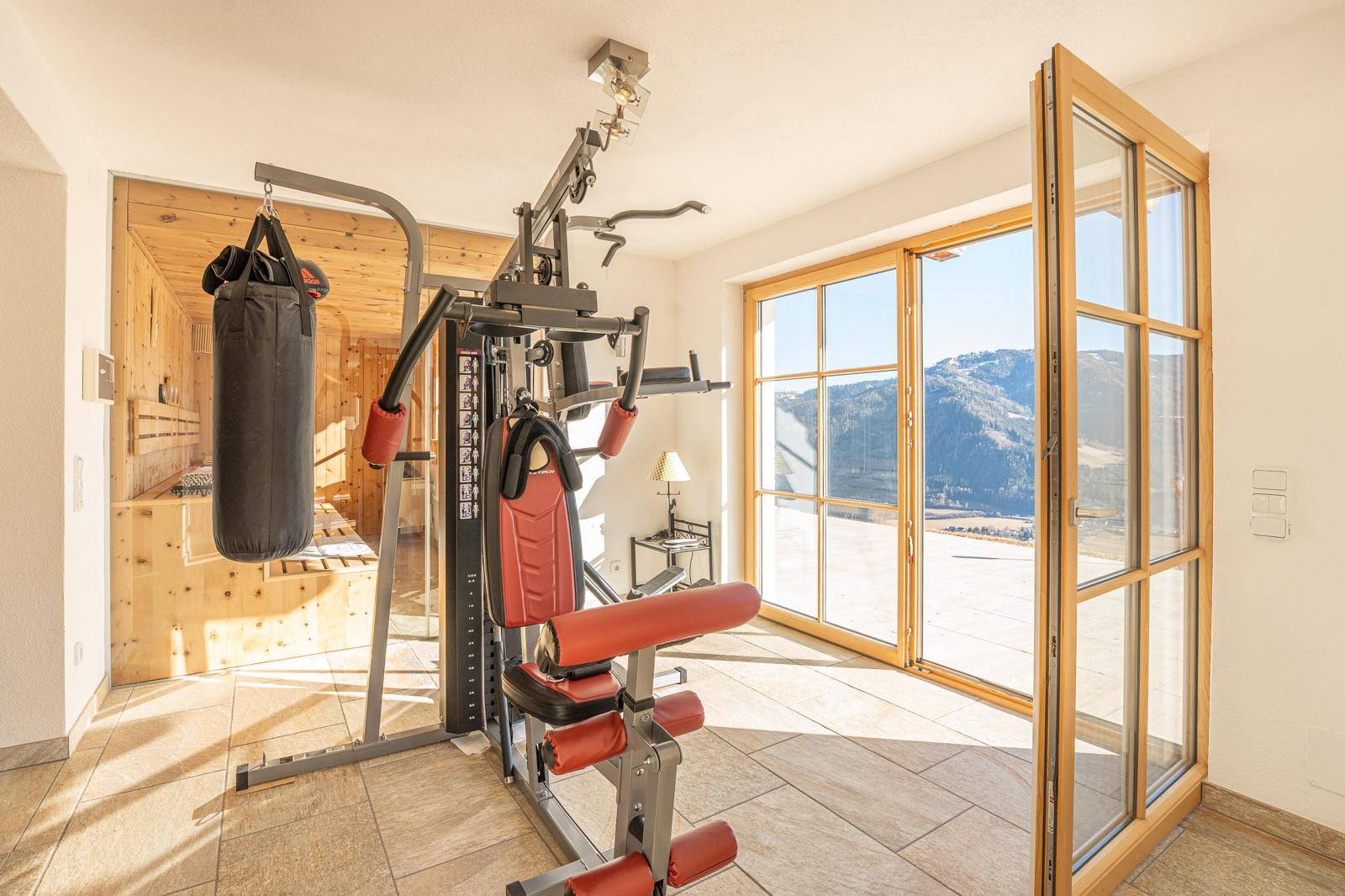 Fitnessraum mit Trainingsgeräten, Sauna im Hintergrund und Zugang zur Terrasse mit Bergblick.