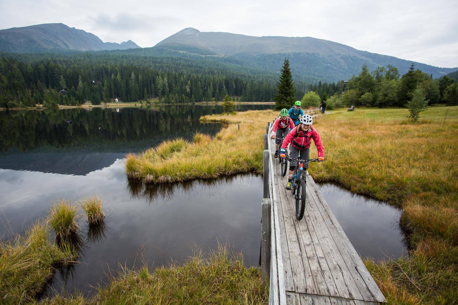 Mountainbiker überqueren eine Holzbrücke über einen See, umgeben von Wald und Bergen.