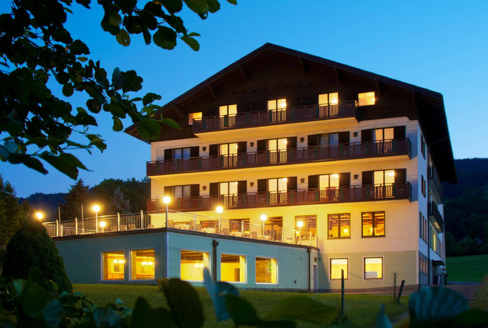 Außenansicht des Gebäudes bei Dämmerung mit beleuchteten Fenstern, Balkonen und einer Terrasse.