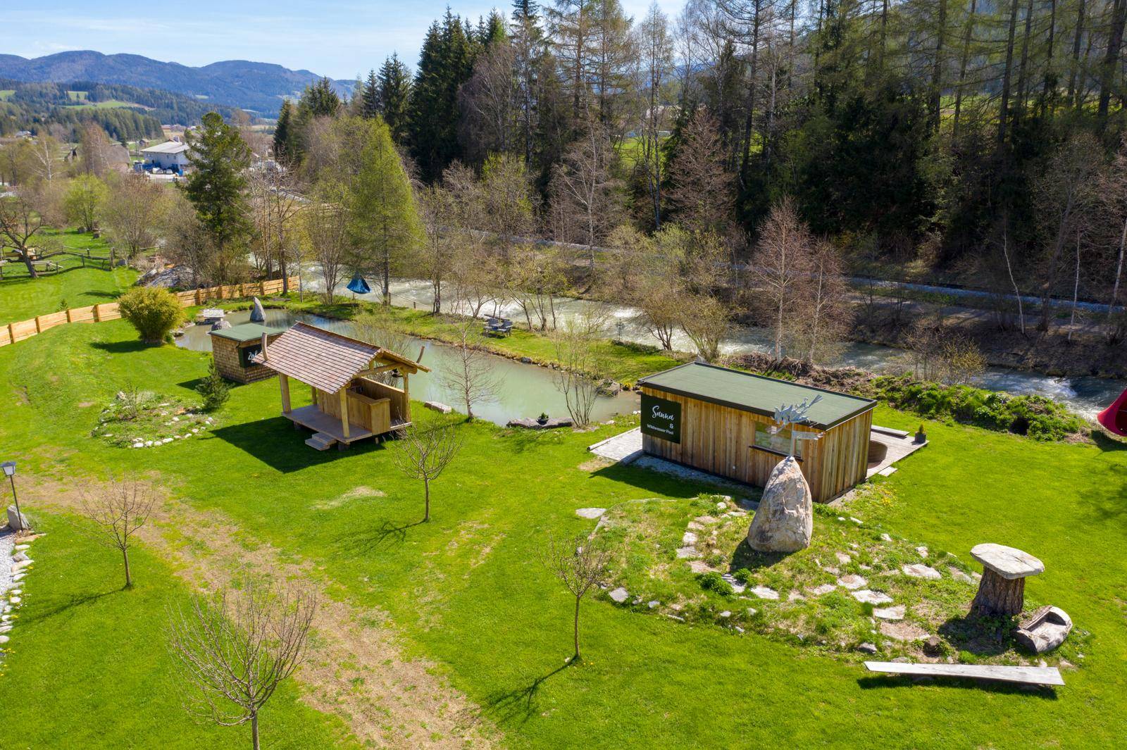Luftaufnahme einer weitläufigen Anlage mit Holzhäusern, grünen Wiesen und Flusslandschaft.
