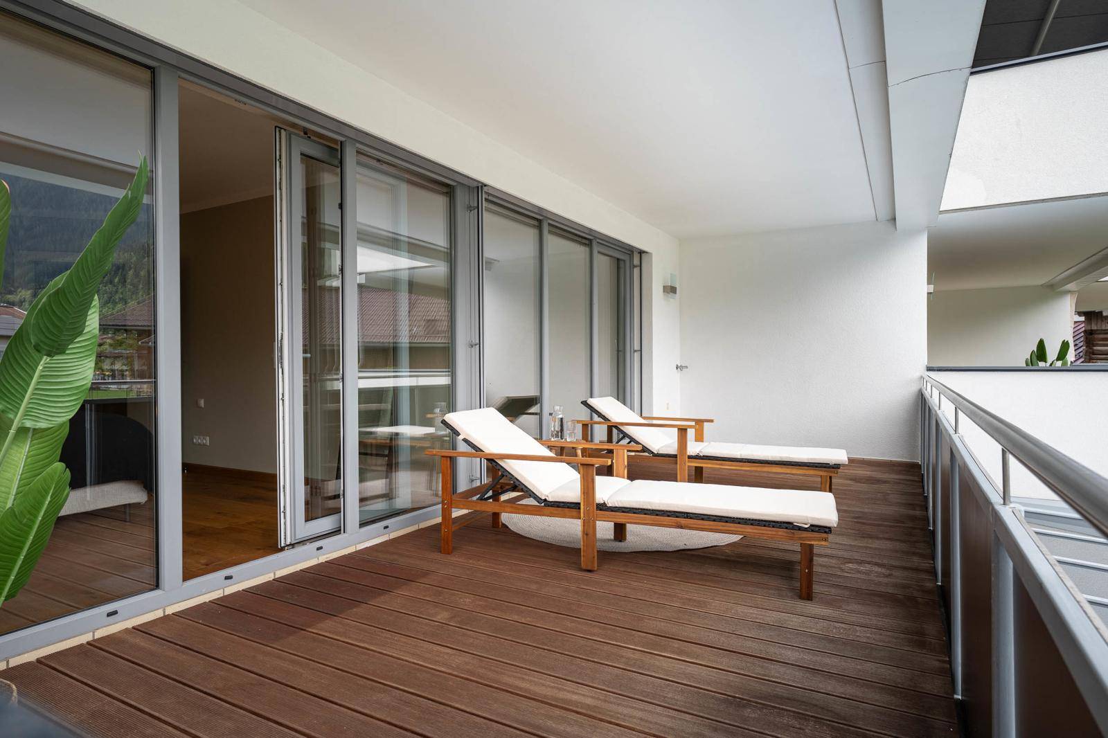 Geräumiger Balkon mit Holzdeck, bequemen Liegestühlen und großen Glasschiebetüren zum Innenbereich.