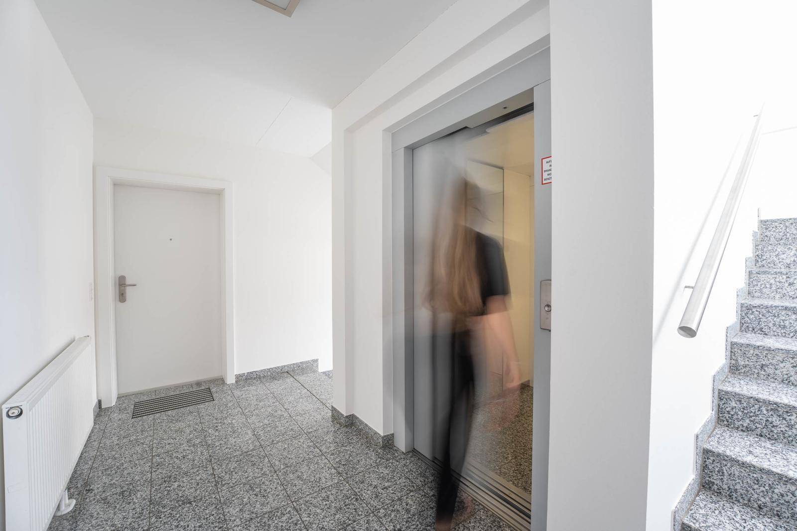 Eingangsbereich mit Aufzug und Treppenhaus, eine Person geht am modernen Aufzug vorbei.