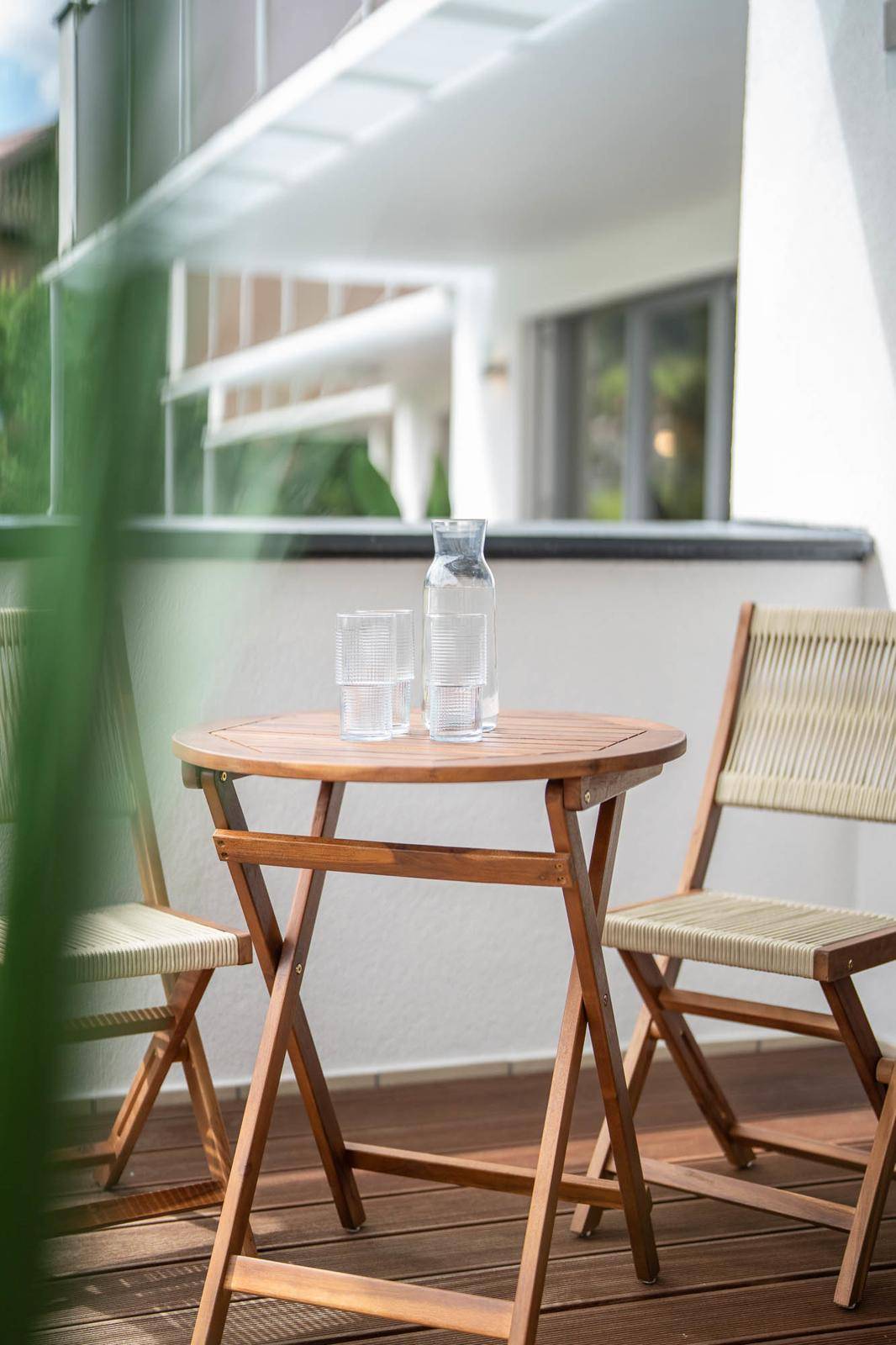 Kleiner Holztisch mit Stühlen auf einem Balkon mit Holzdeck und grüner Bepflanzung im Vordergrund.