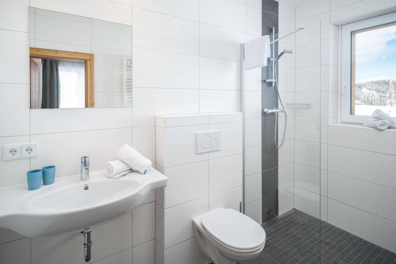 Modernes Badezimmer mit weißen Fliesen, Dusche und Fenster mit Blick auf die Winterlandschaft.