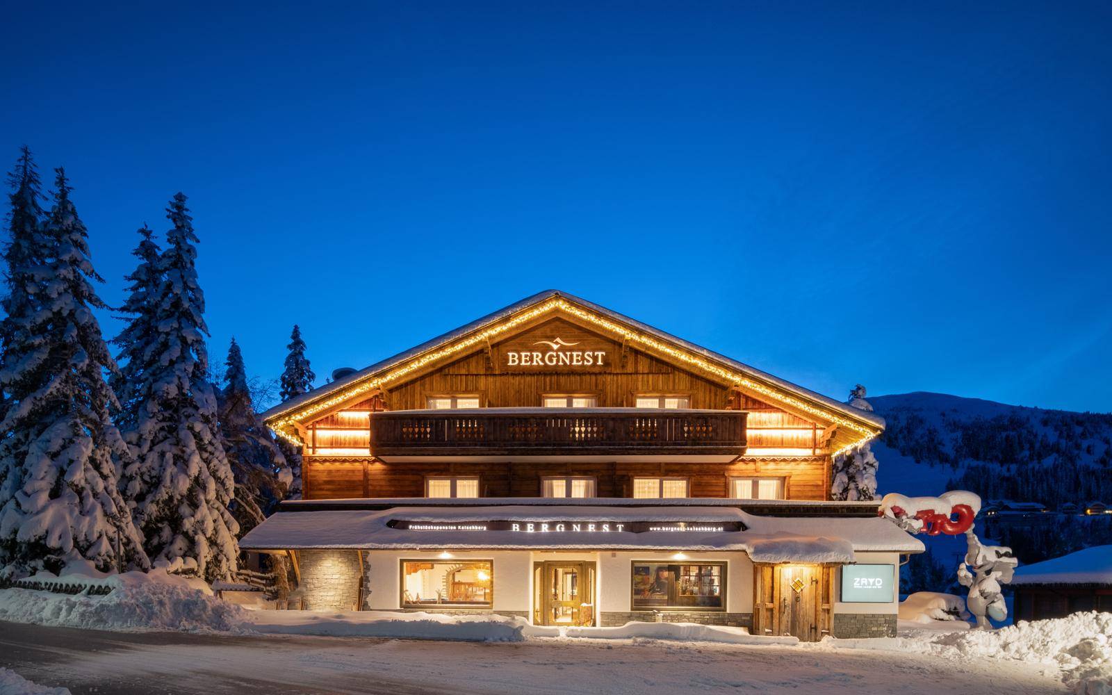 Das beleuchtete Holzchalet in der Dämmerung, umgeben von schneebedeckten Bäumen und einer winterlichen Berglandschaft.
