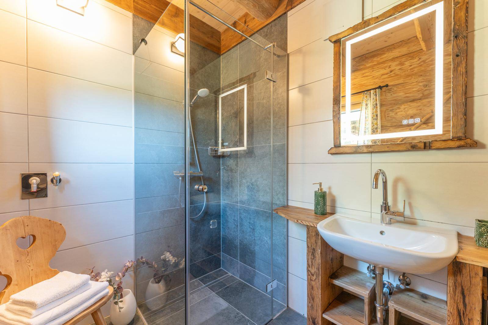 Helles Badezimmer mit Glasdusche, beleuchtetem Spiegel und Holzwaschtisch.