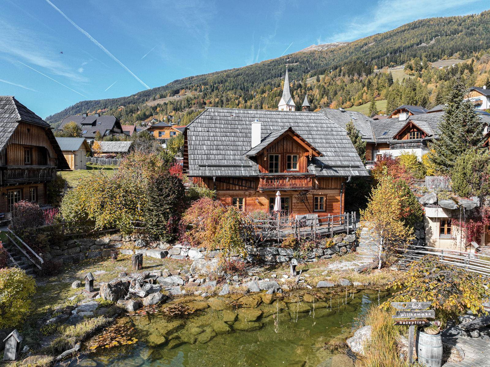 Charmantes Holzhaus in alpiner Umgebung mit Teich und Blick auf die umliegenden Berge und das Dorf.