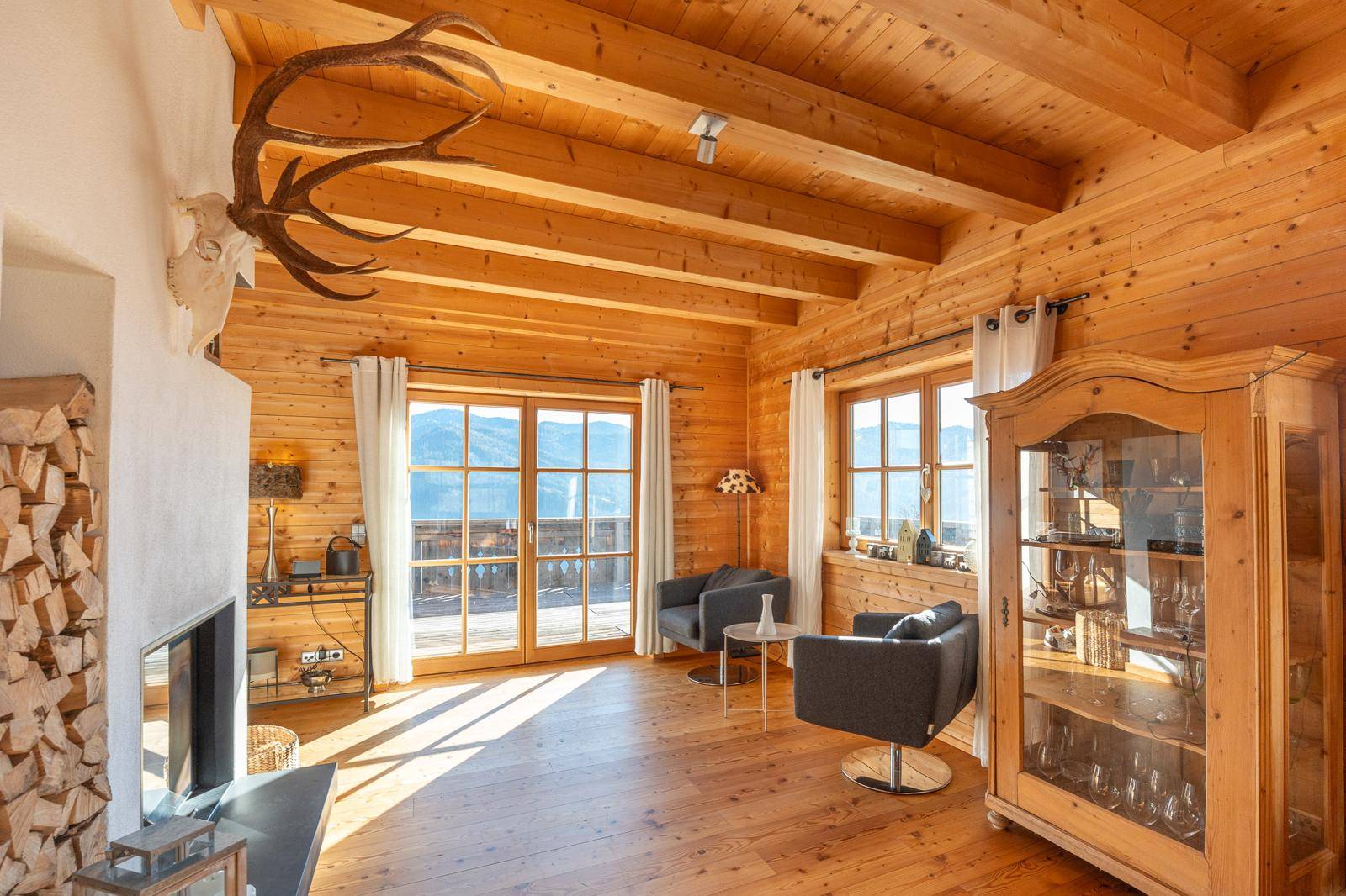 Lichtdurchflutetes Wohnzimmer mit Kamin, Holzwänden, Holzbalkendecke und Zugang zum Balkon.