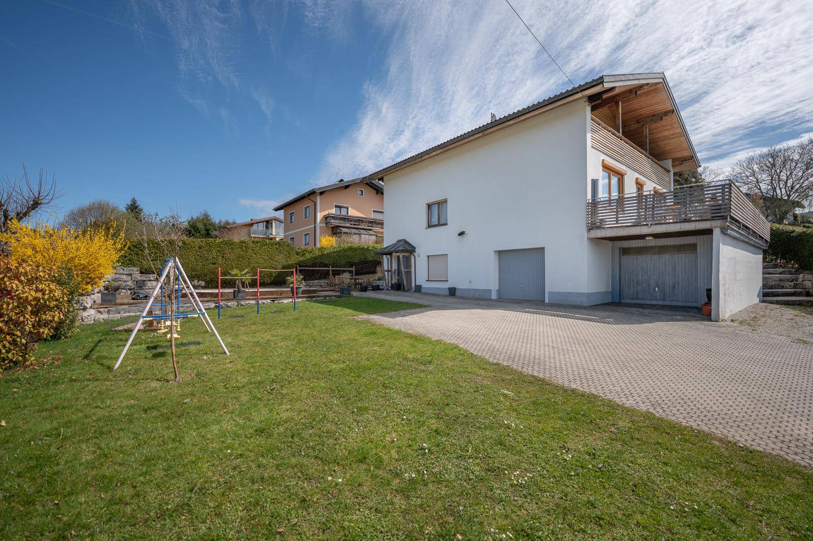 Seitenansicht des Hauses mit gepflegtem Rasen, Spielgeräten und großzügiger Auffahrt, umgeben von Grün.