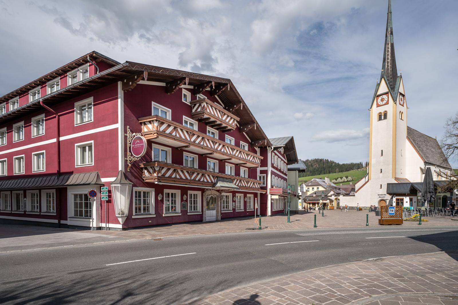 Traditionelles rotes Gebäude mit Holzbalkonen und einer Kirche im Hintergrund an einem sonnigen Tag.