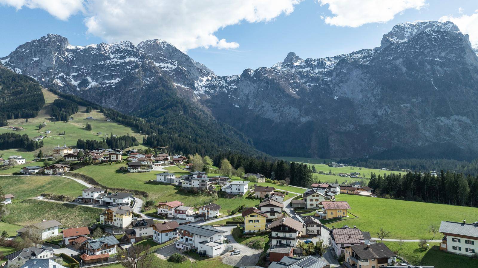 Weitläufige Panoramaansicht eines Dorfes in einem Tal, umgeben von grünen Hügeln und schneebedeckten Bergen.