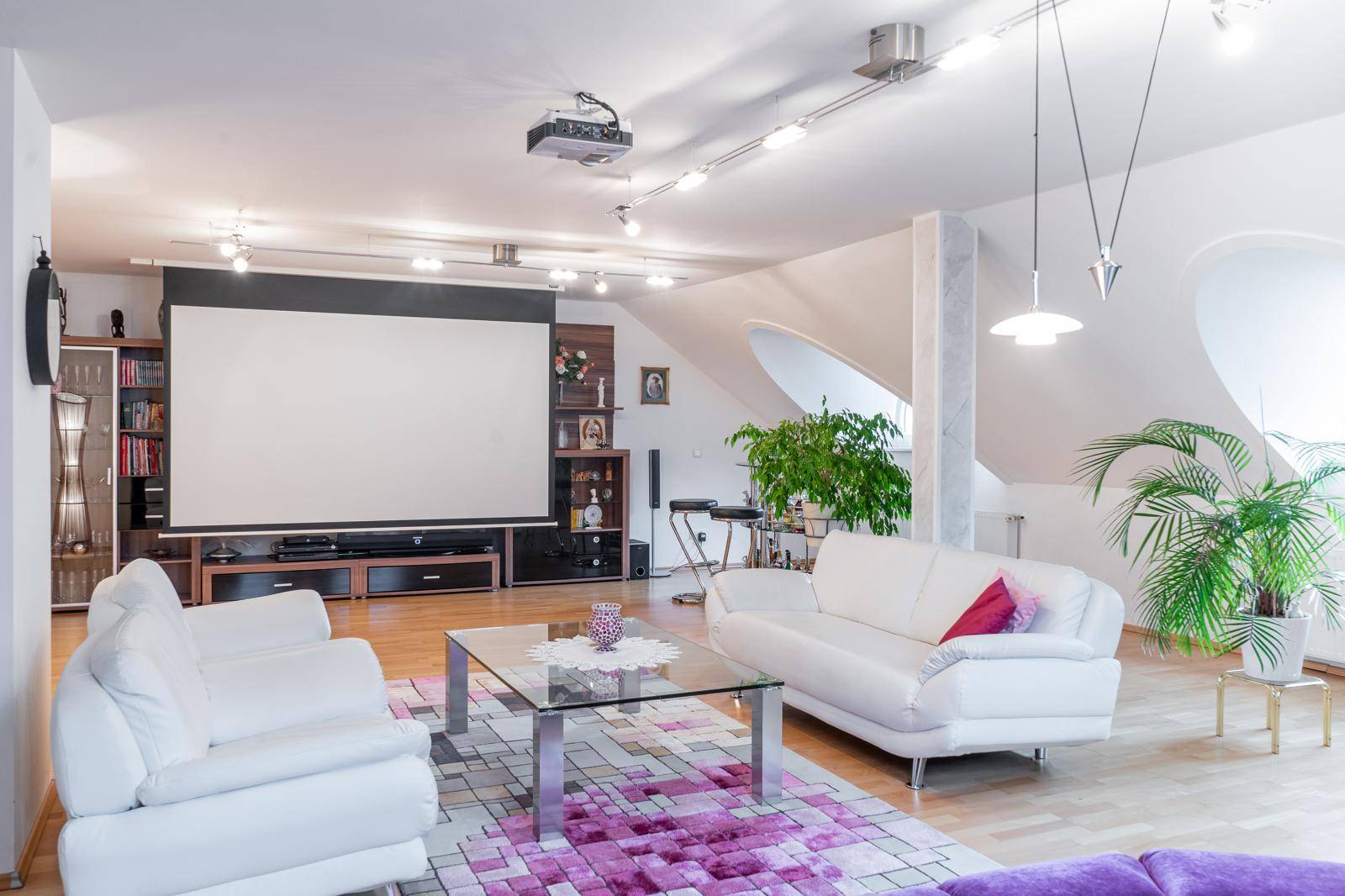 Geräumiges Wohnzimmer mit großer Leinwand, weißen Sofas und modernen Lichtschienen.