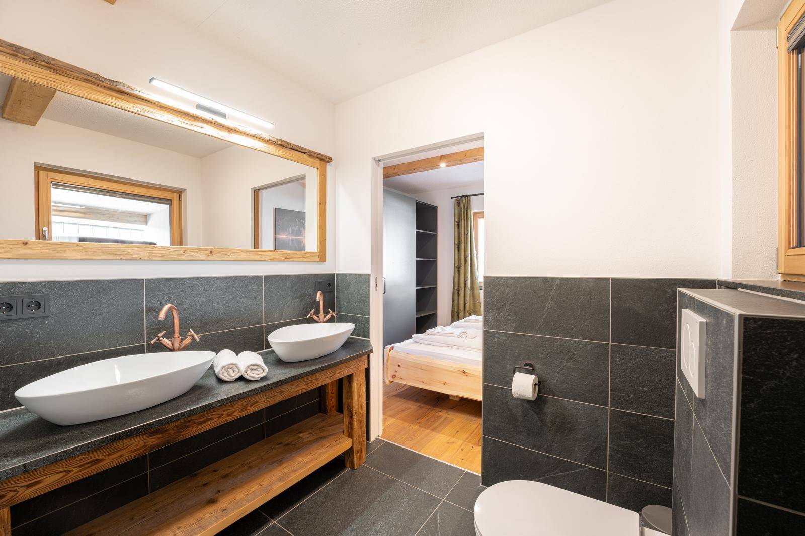 Modernes Badezimmer mit Doppelwaschbecken, dunklen Fliesen und Zugang zum Schlafzimmer.