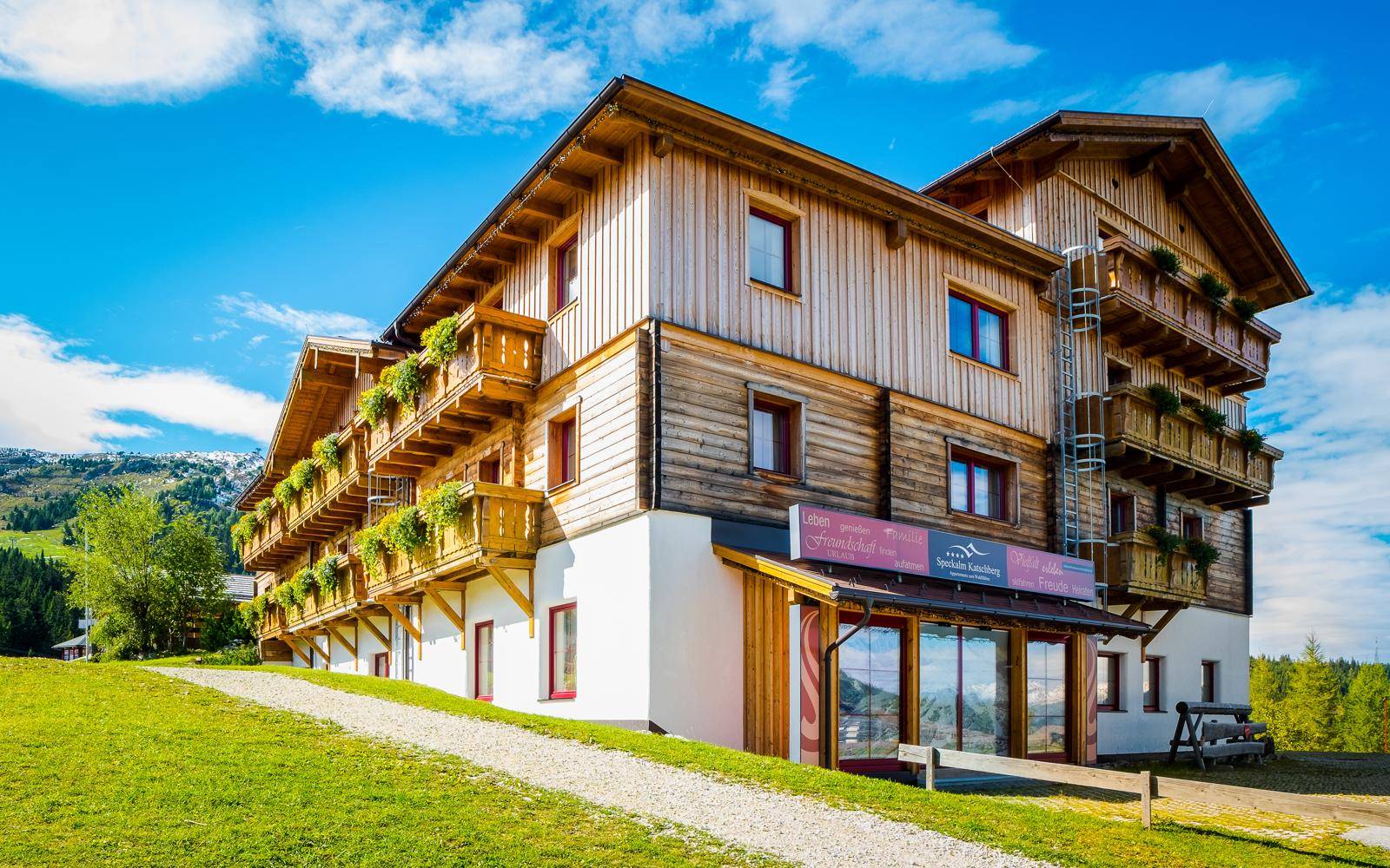 Traditionelles Holzgebäude im alpinen Stil, umgeben von grünen Wiesen unter blauem Himmel.