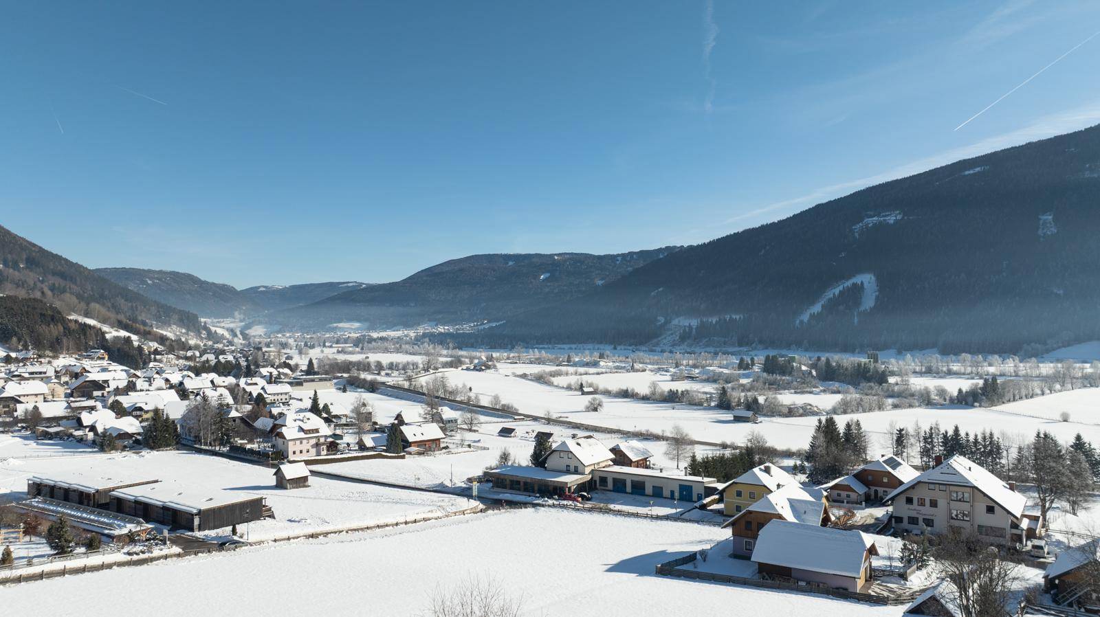Weitläufiger Blick über ein verschneites Tal mit einem Dorf, Feldern und bewaldeten Bergen im Hintergrund.