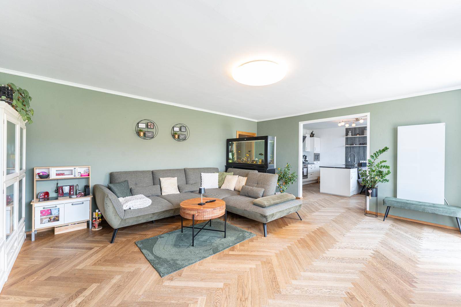 Geräumiges Wohnzimmer mit Fischgrätparkett, großem Sofa und Blick in die offene Küche, ideal für Familien.