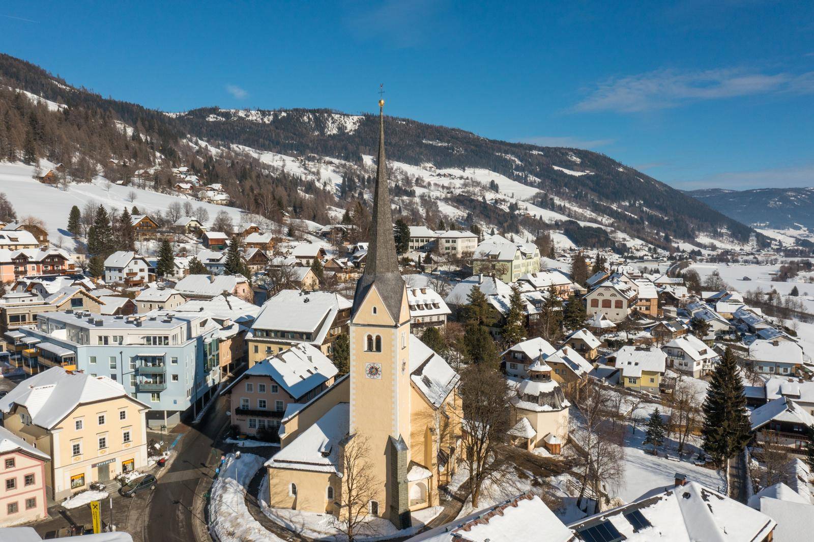 Eine verschneite Dorfansicht mit einer markanten Kirche im Zentrum, umgeben von Winterlandschaft.