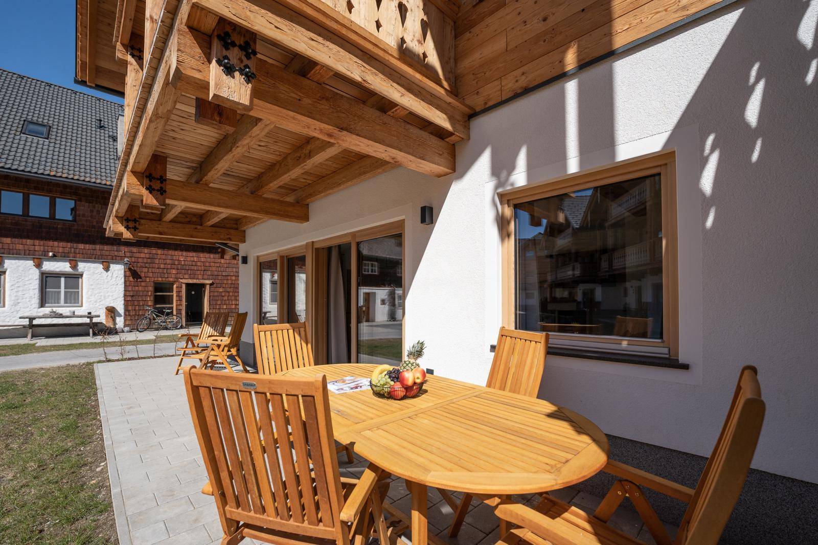 Gemütliche Terrasse mit Holztisch und Stühlen, umgeben von Grünflächen und dem Gebäude.