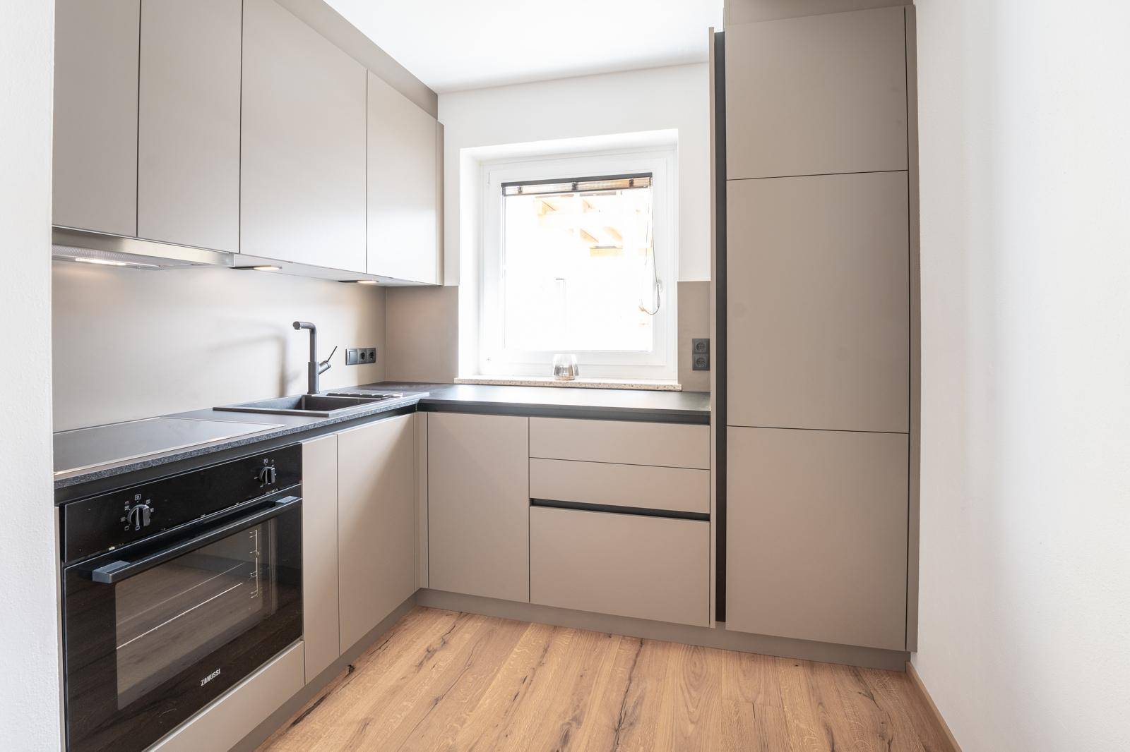 Moderne Einbauküche in Beige mit dunkler Arbeitsplatte und Fenster für Tageslicht.