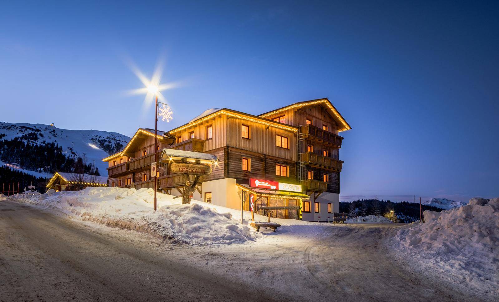Beleuchtetes Holzgebäude im alpinen Stil bei Nacht, umgeben von Schnee und Bergen.