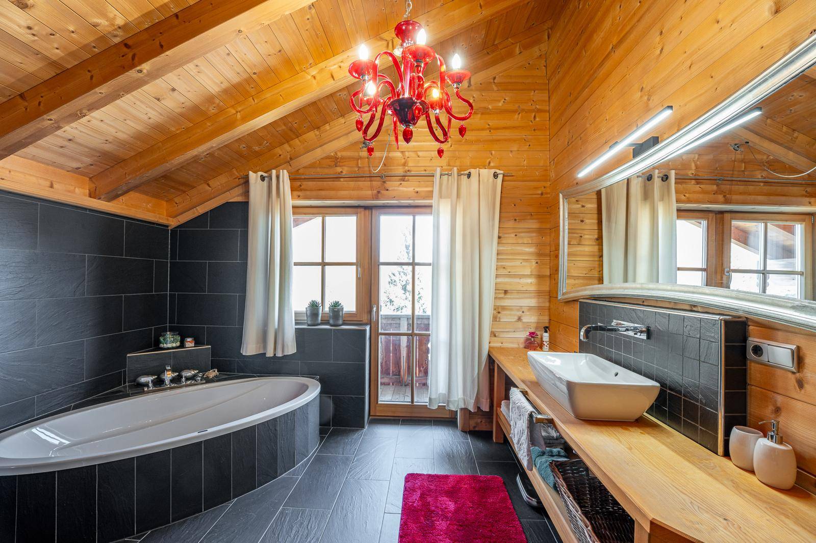 Modernes Badezimmer mit Badewanne, dunklen Fliesen, Holzwänden und rotem Kronleuchter.