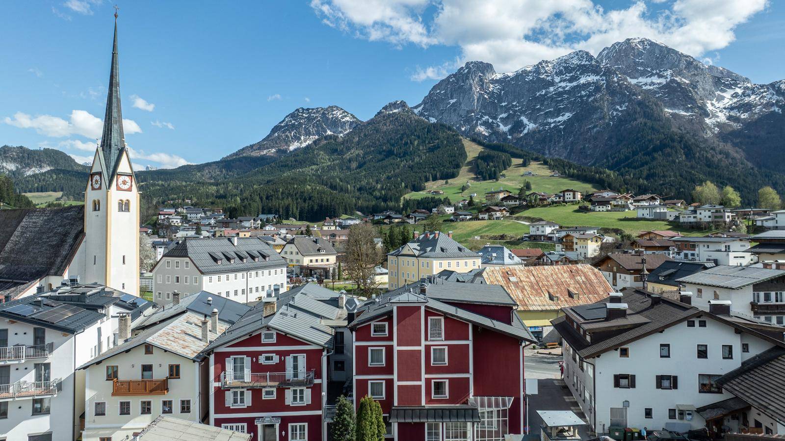 Panoramablick auf ein malerisches Dorf mit Kirche und schneebedeckten Bergen im Hintergrund unter blauem Himmel.