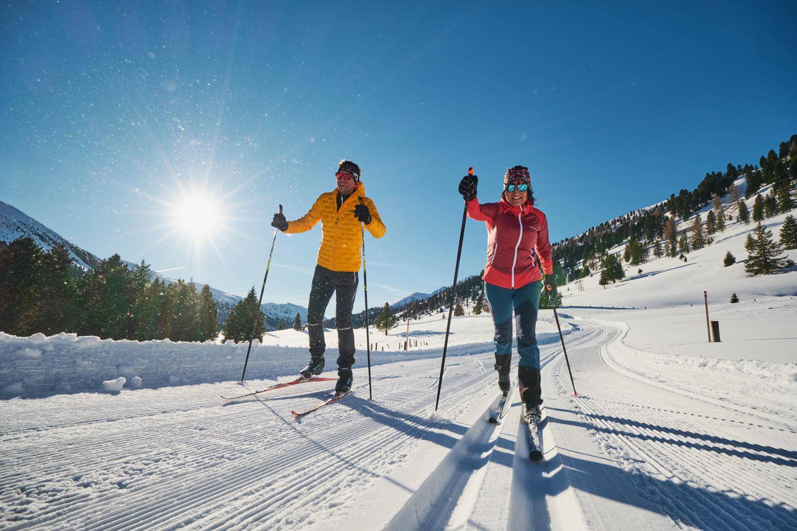Zwei Personen beim Langlaufen auf einer präparierten Loipe in einer sonnigen Winterlandschaft.