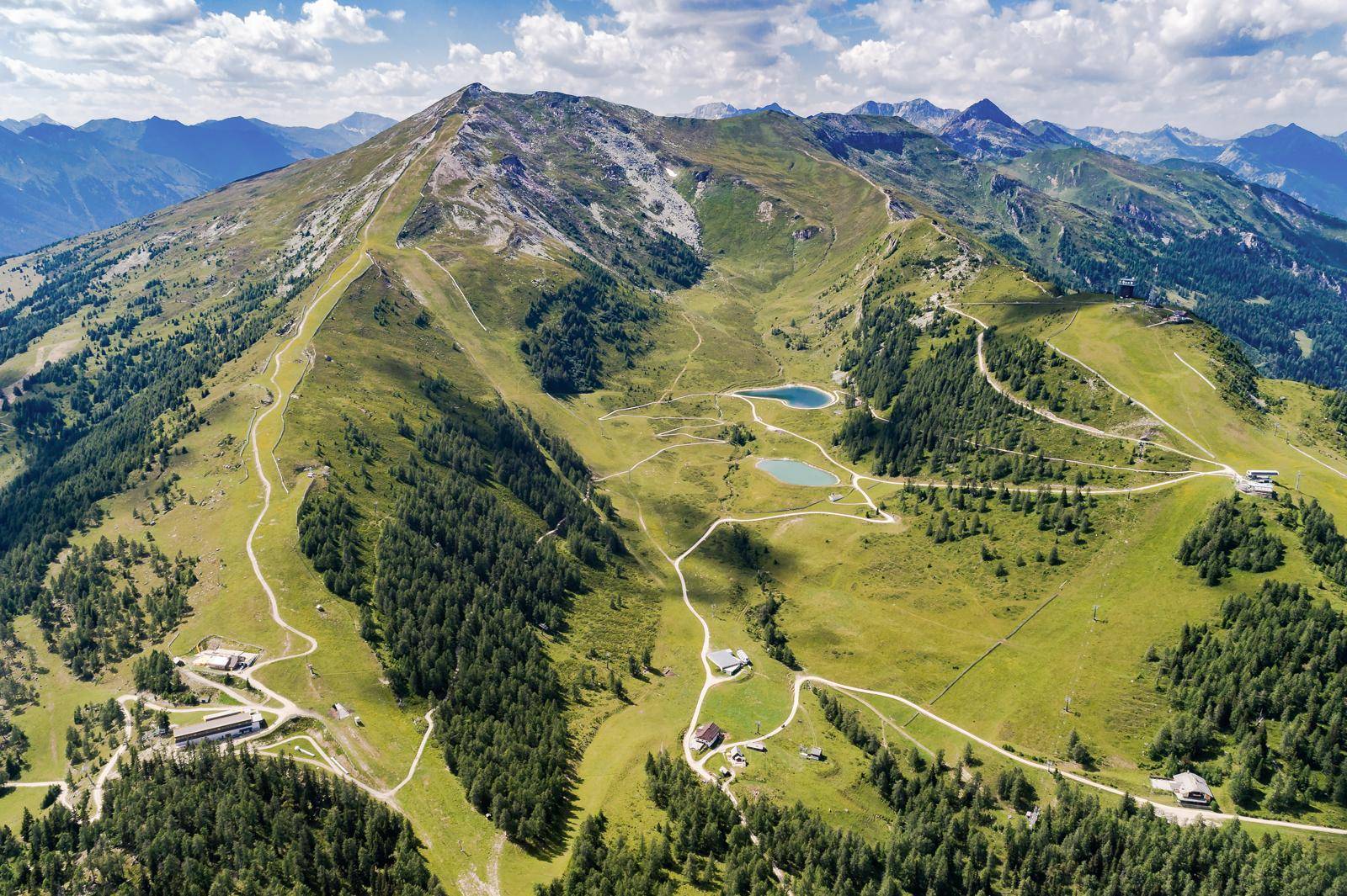Eine weite Luftaufnahme einer grünen Berglandschaft mit Wäldern, Wanderwegen und kleinen Seen.