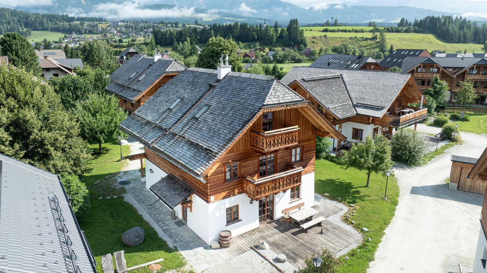 Luftaufnahme eines traditionellen Holzhauses mit Schindeldach, Balkonen und einer Holzterrasse in alpiner Landschaft.