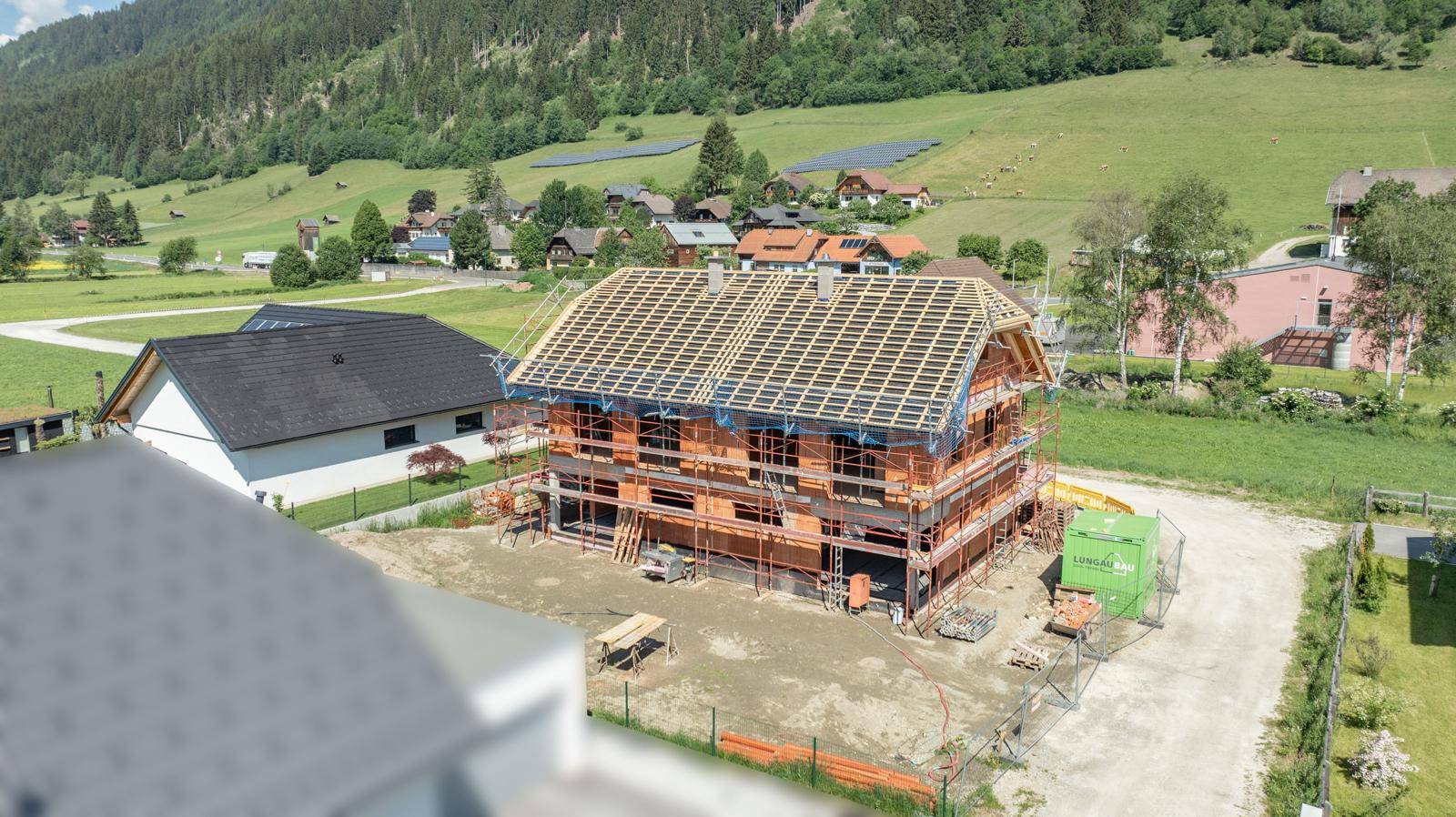 Neubau im Rohbau mit Dachstuhl, umgeben von anderen Häusern und grünen Hügeln.