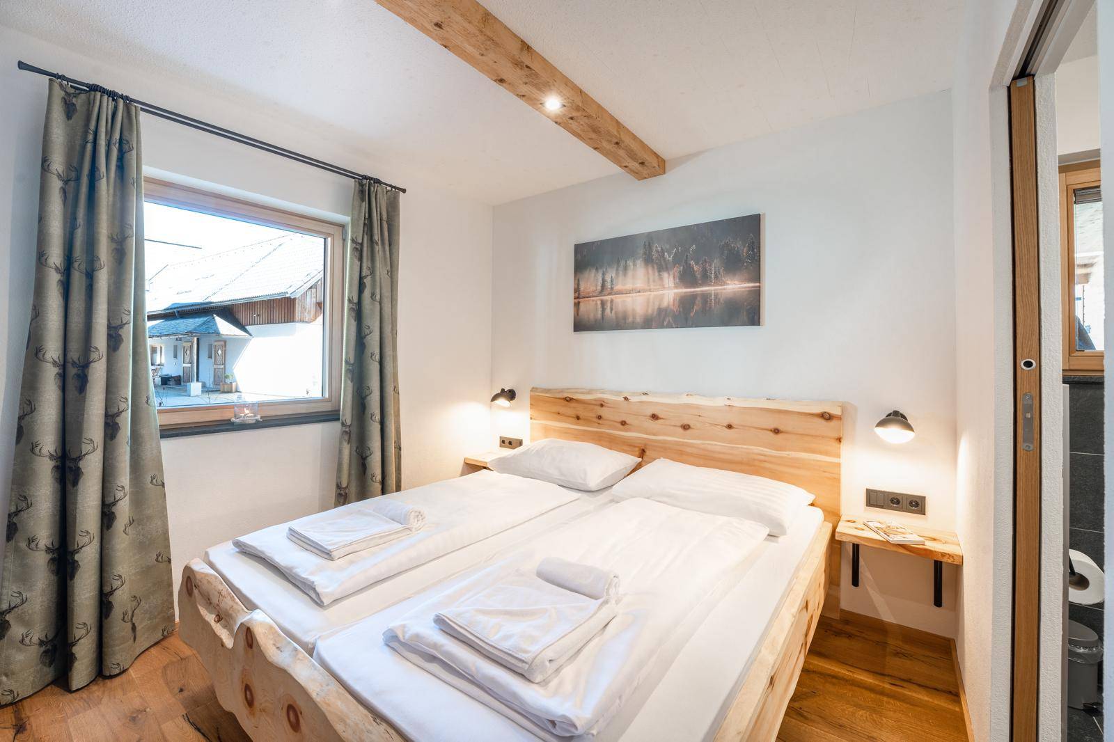 Helles Schlafzimmer mit Holzbett, Holzbalken und Fenster mit Blick nach draußen.
