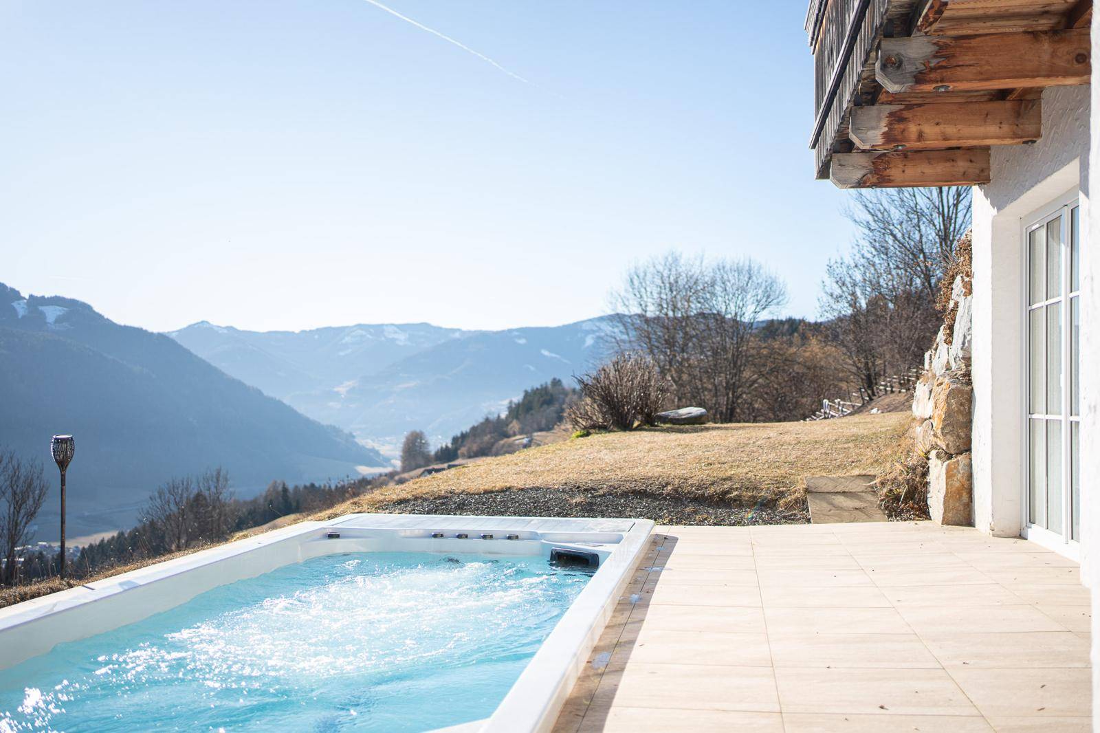 Sonniger Außenbereich mit Whirlpool und Fliesenboden, umgeben von einer malerischen Berglandschaft.