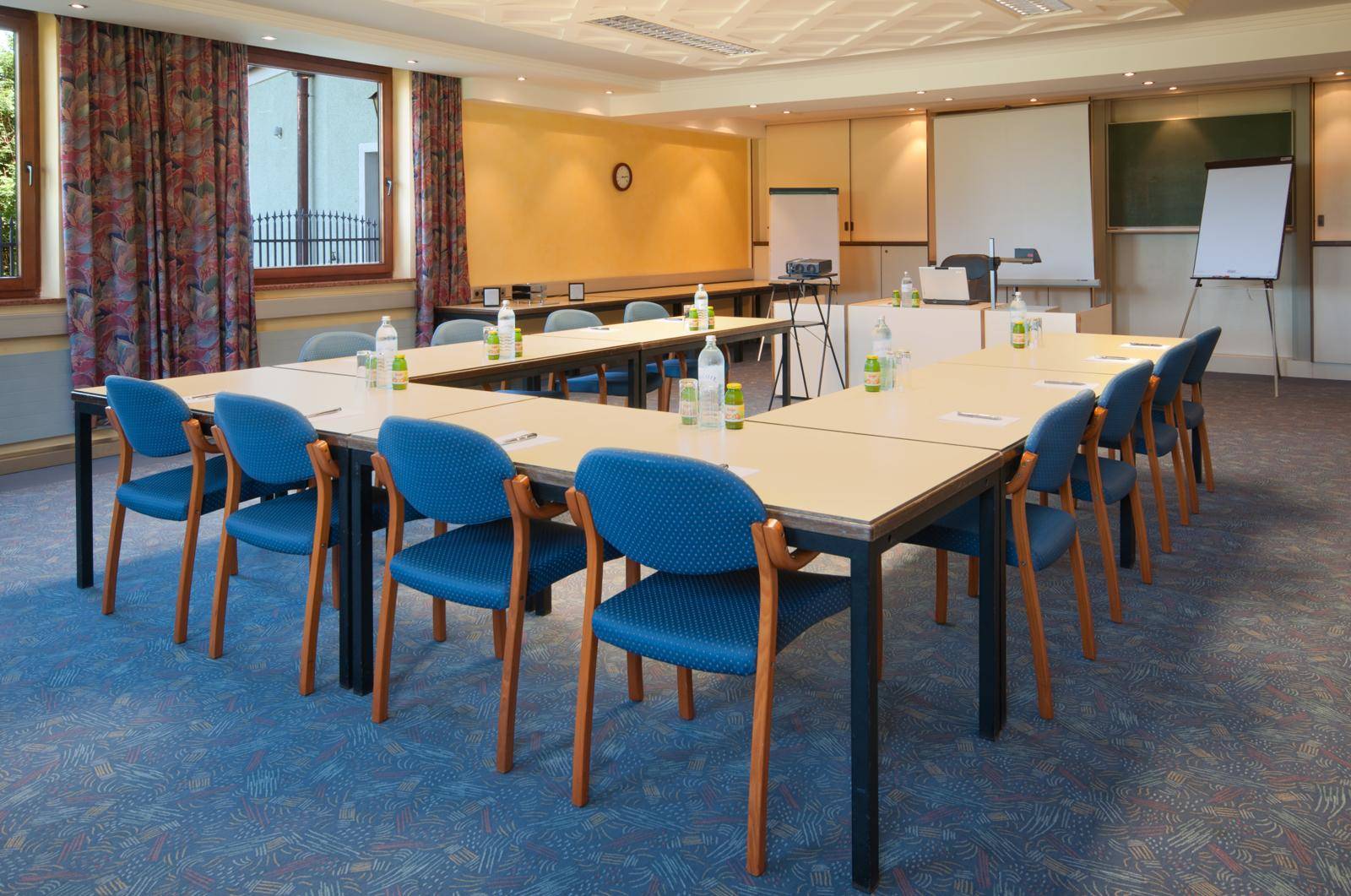 Heller Konferenzraum mit blauen Stühlen, Tischen in U-Form und Blick ins Grüne.